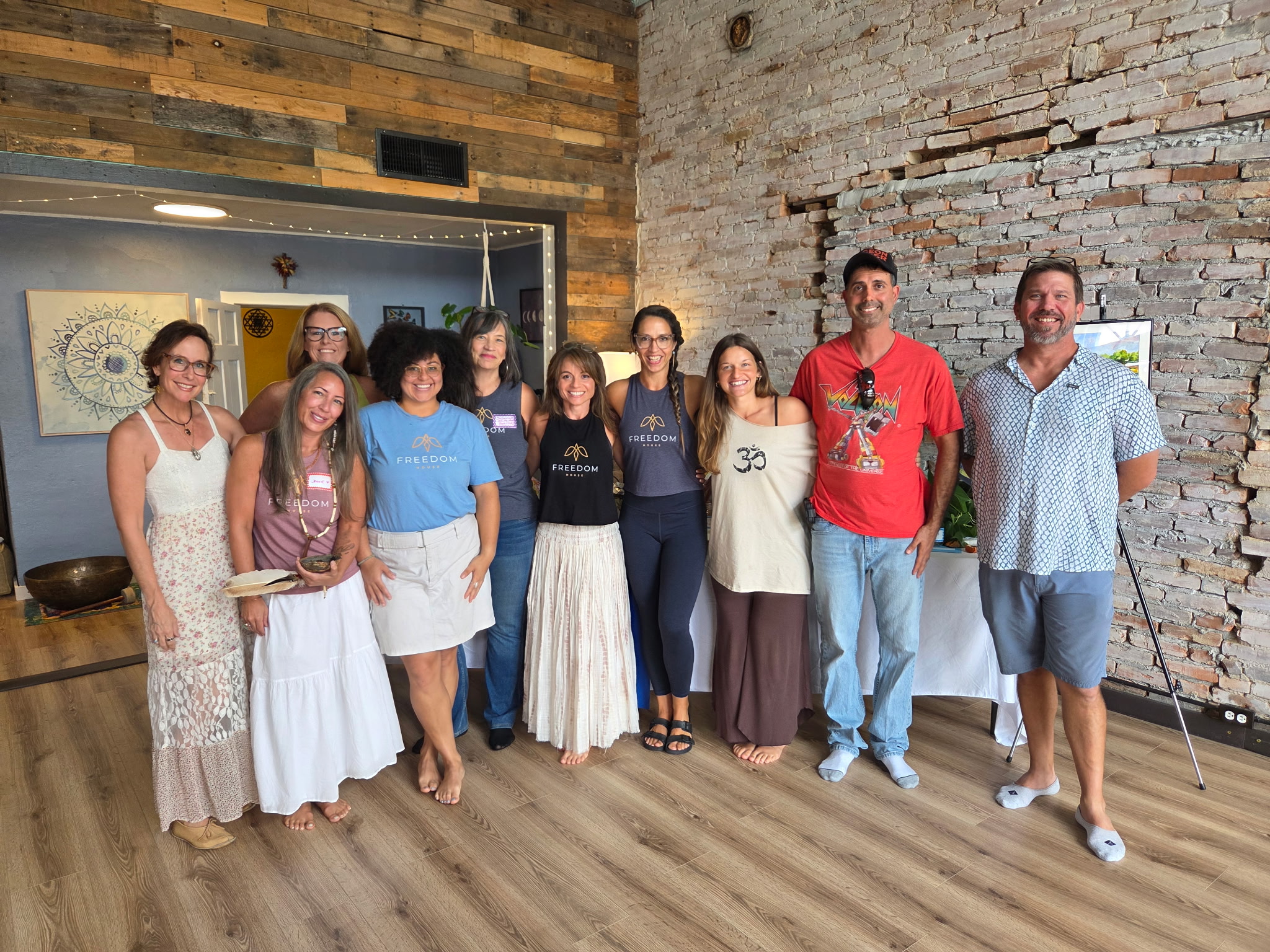 Group of ten people standing indoors against a brick wall, smiling, with wooden flooring and a window in the background.