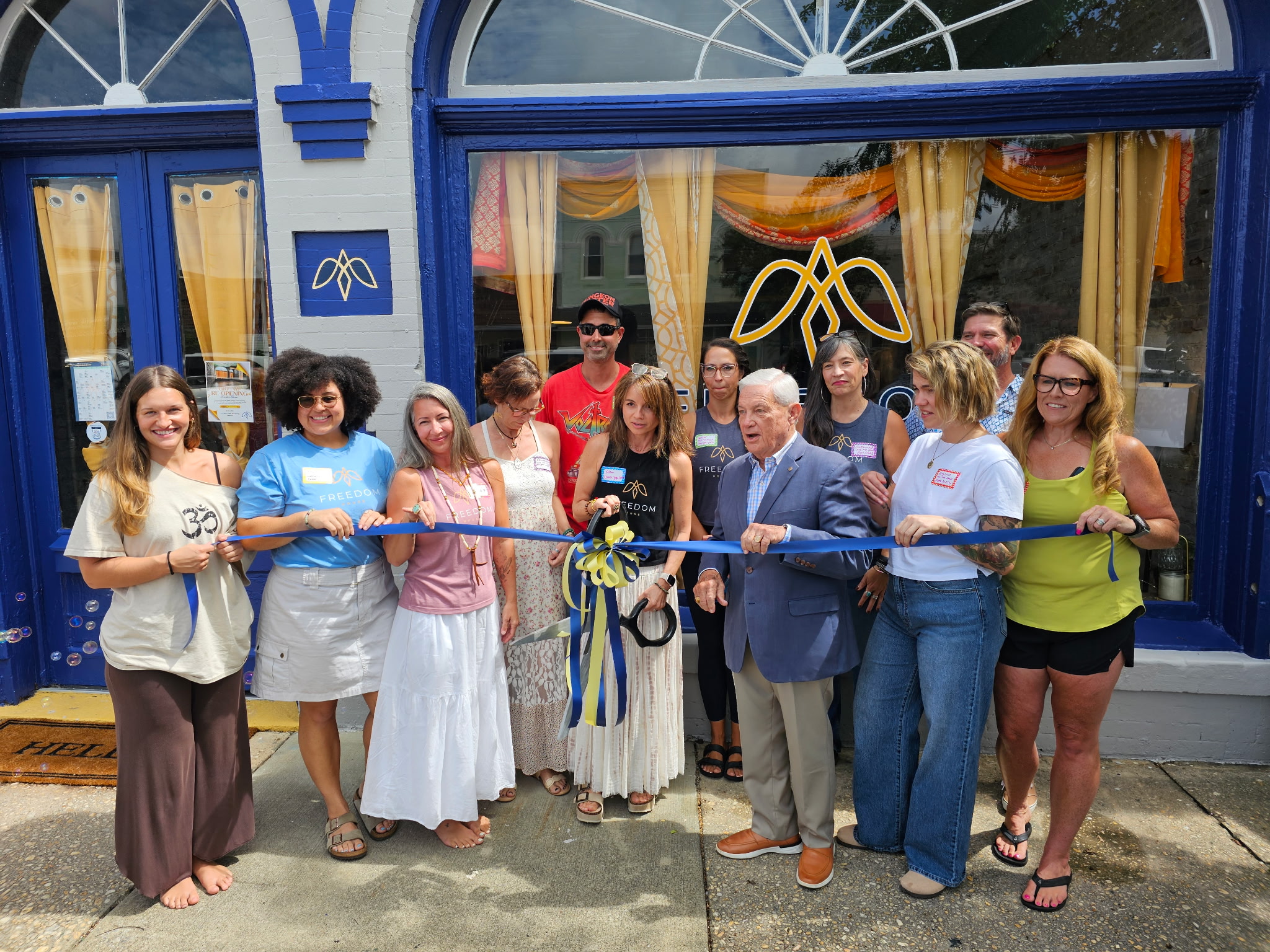 Group of people participating in a ribbon-cutting event outside a building with blue and yellow decor.