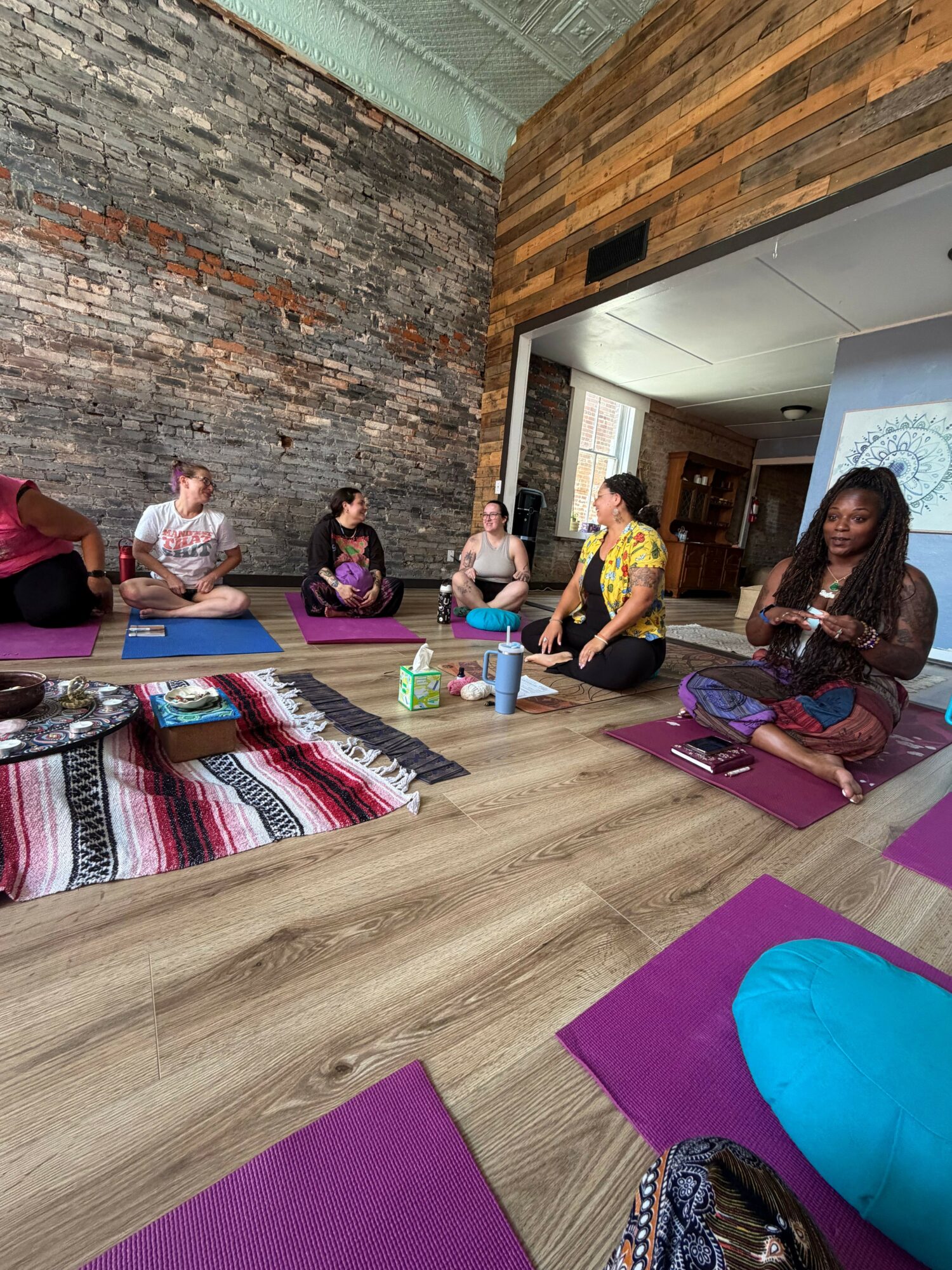 People sitting on yoga mats in a room with a brick wall, some holding books or papers, and a wooden wall on the left.