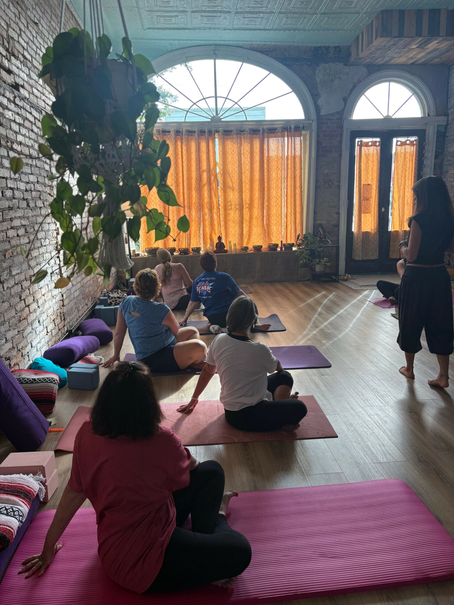 People practicing yoga on mats in a spacious room with large arched windows and orange curtains.