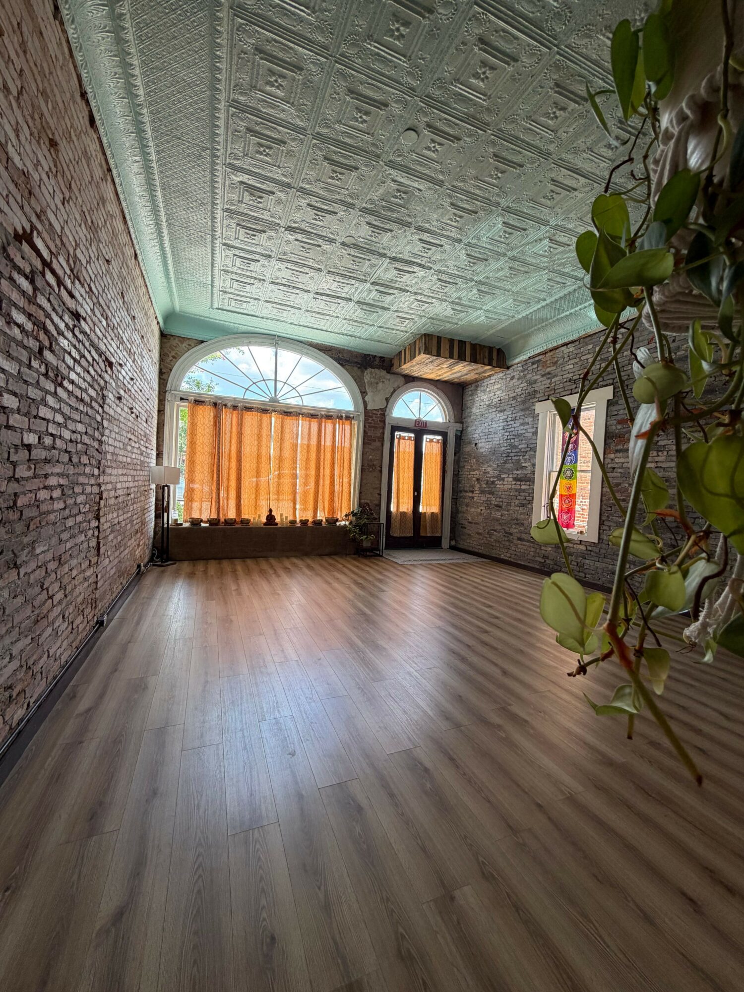 Room with a large arched window, wooden floor, brick wall, and potted plant, viewed from an angle.