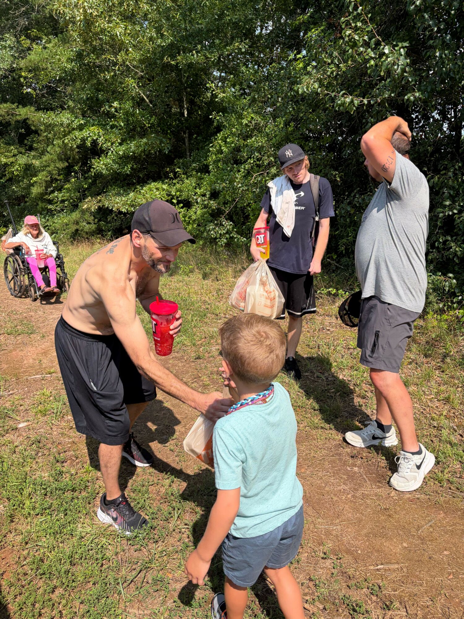 Woman crouching and shaking hands with a young boy outdoors, with two other people in the background, trees, and grass.