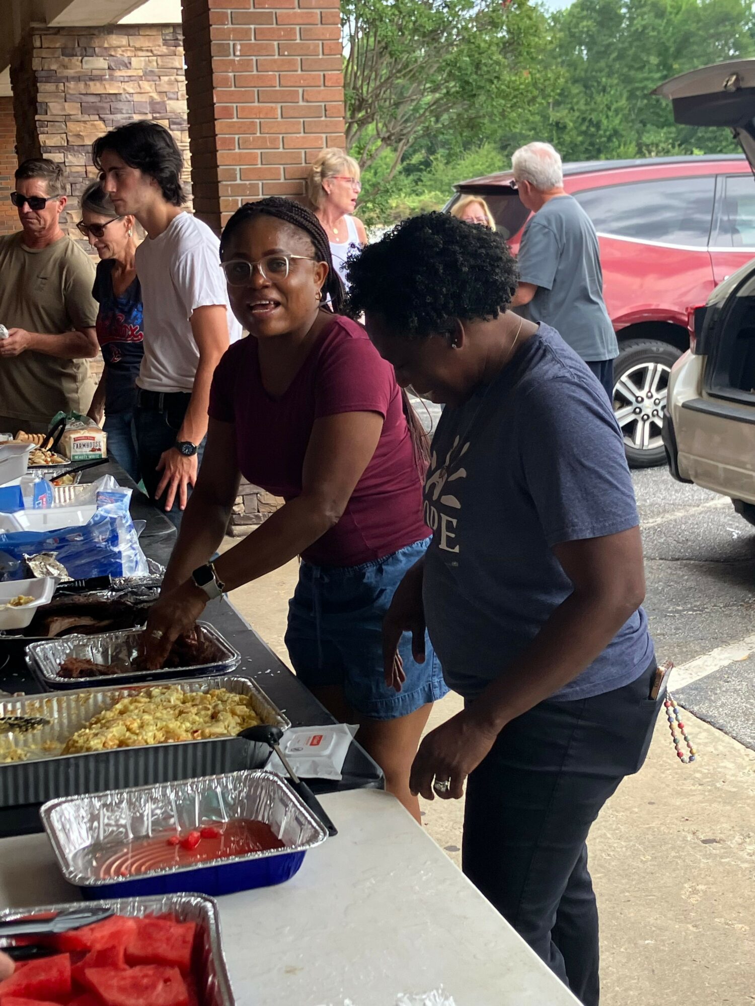 Two people preparing food outdoors, with others in the background near a brick wall and parked cars.
