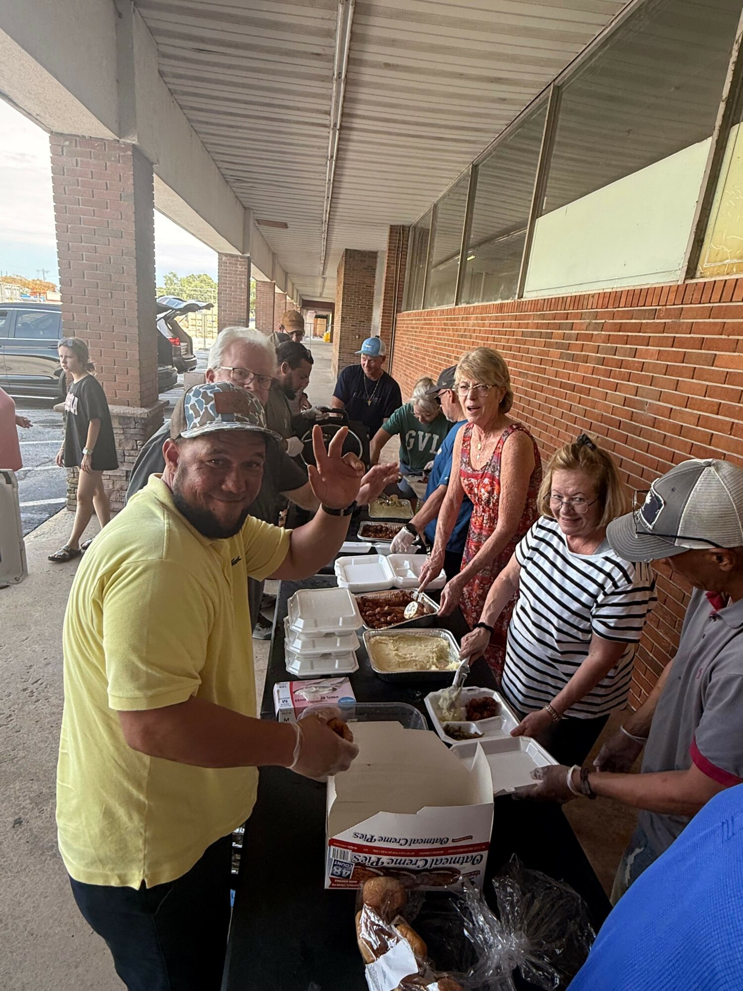 People gather around tables with food outside a brick building, some serving and others waiting in line.