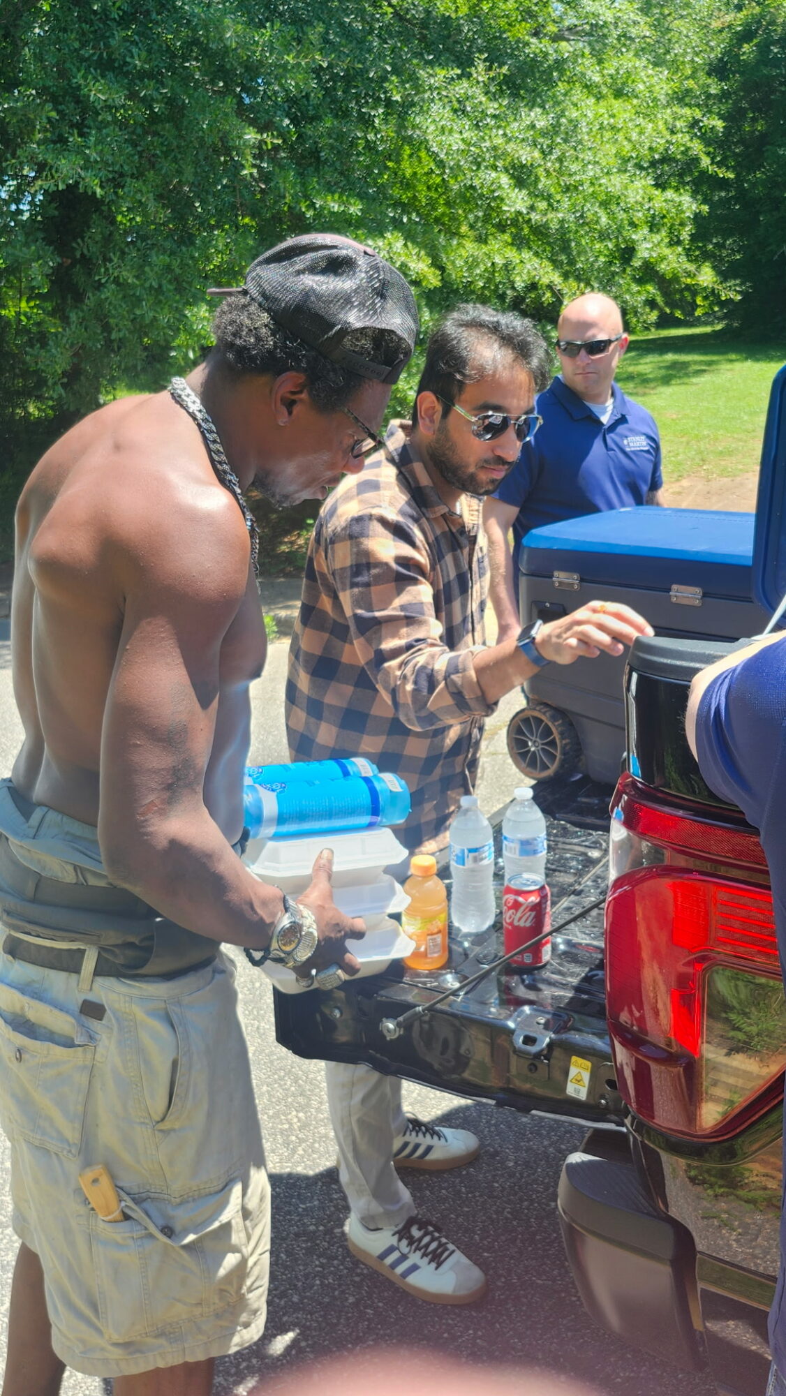 Three men outdoors near a car, one shirtless holding a bottle, another in a plaid shirt, and a third in sunglasses in background.