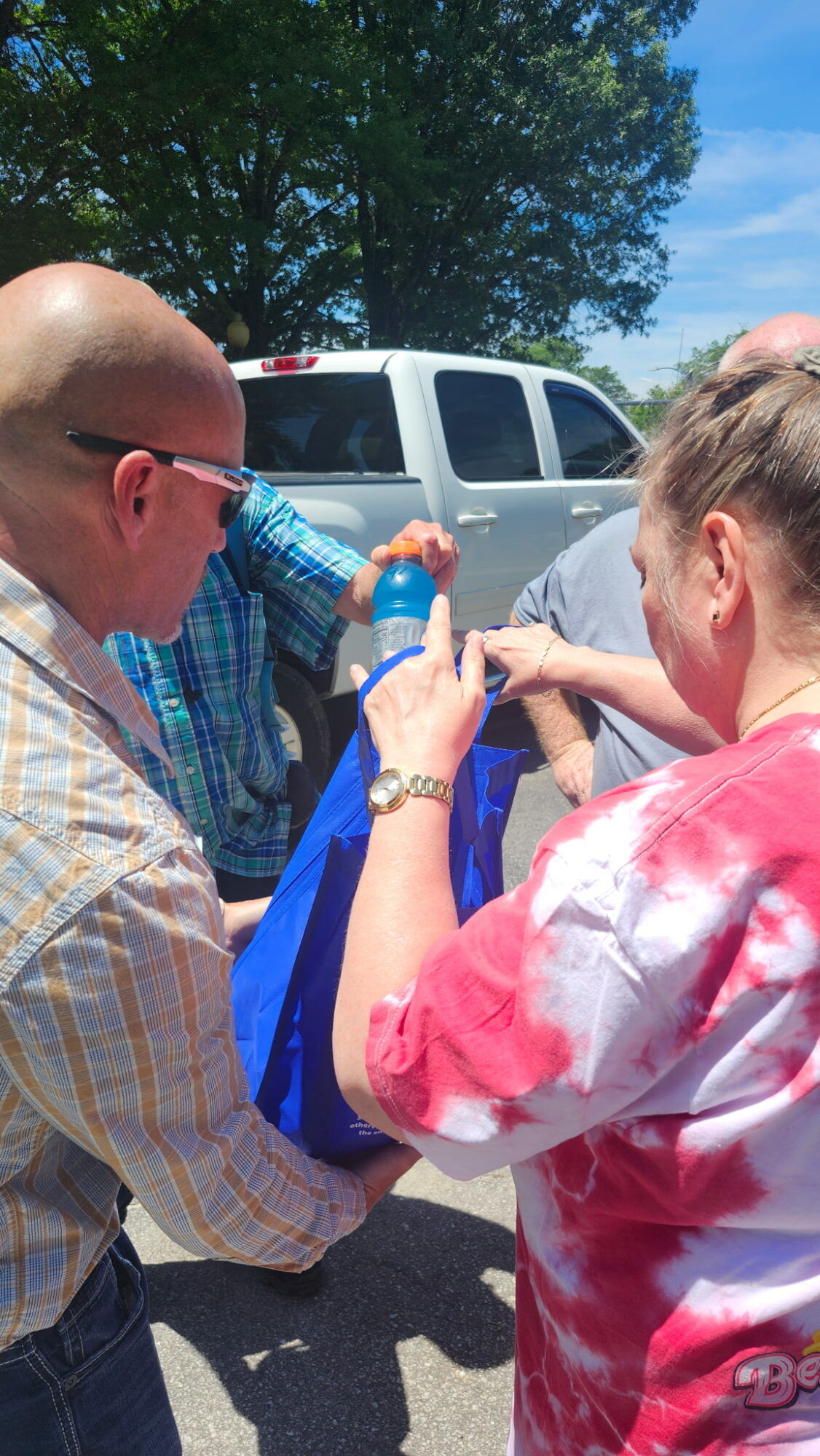 Three people outdoors near a white vehicle, with trees and blue sky in background, engaging with a blue bag.