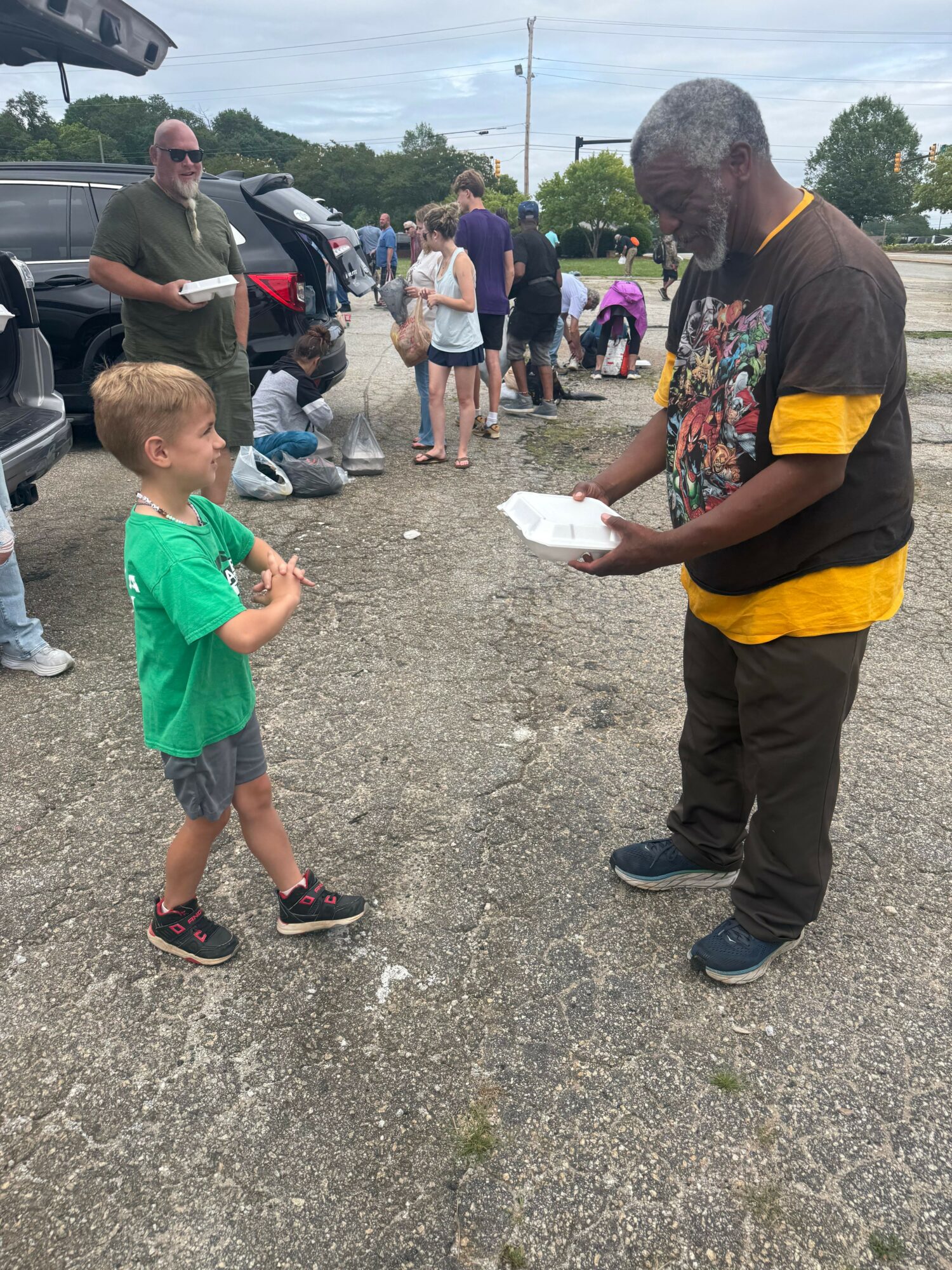 A man in black and yellow clothing interacts with a young boy in a green shirt on a gravel surface, with people in the background.