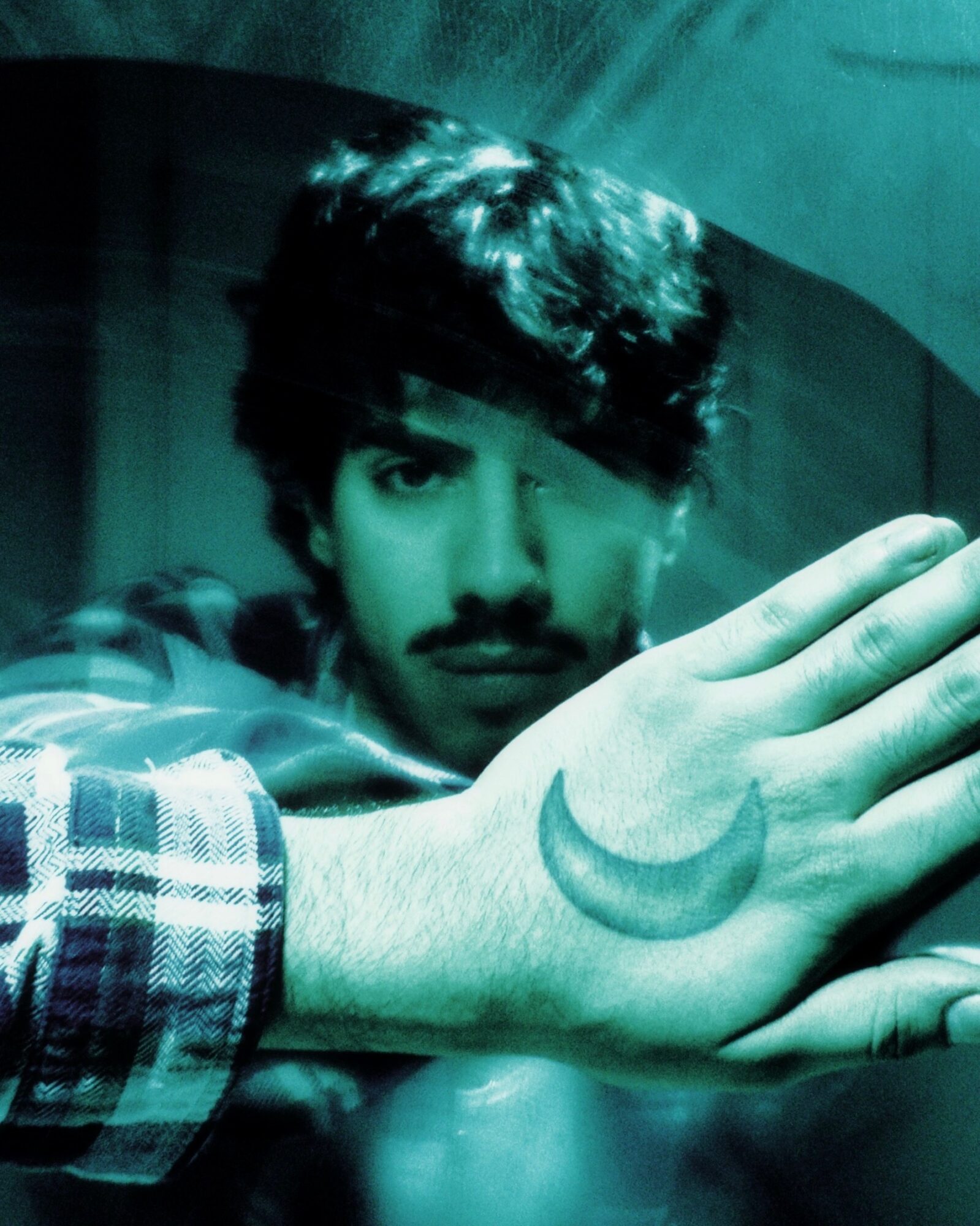 Young man with dark hair and mustache, resting his hand with a crescent moon tattoo, in a dimly lit setting.