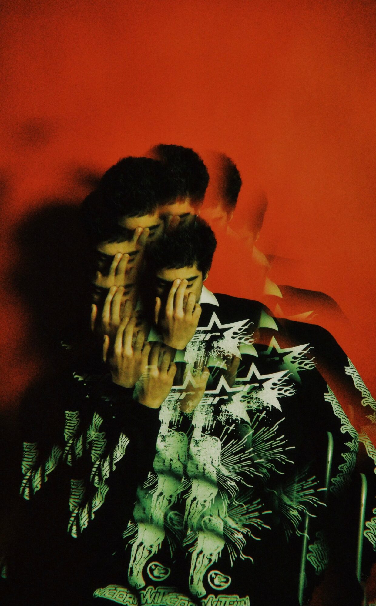 Person covering face with hand, background is red-orange, wearing a patterned shirt with green and white designs.