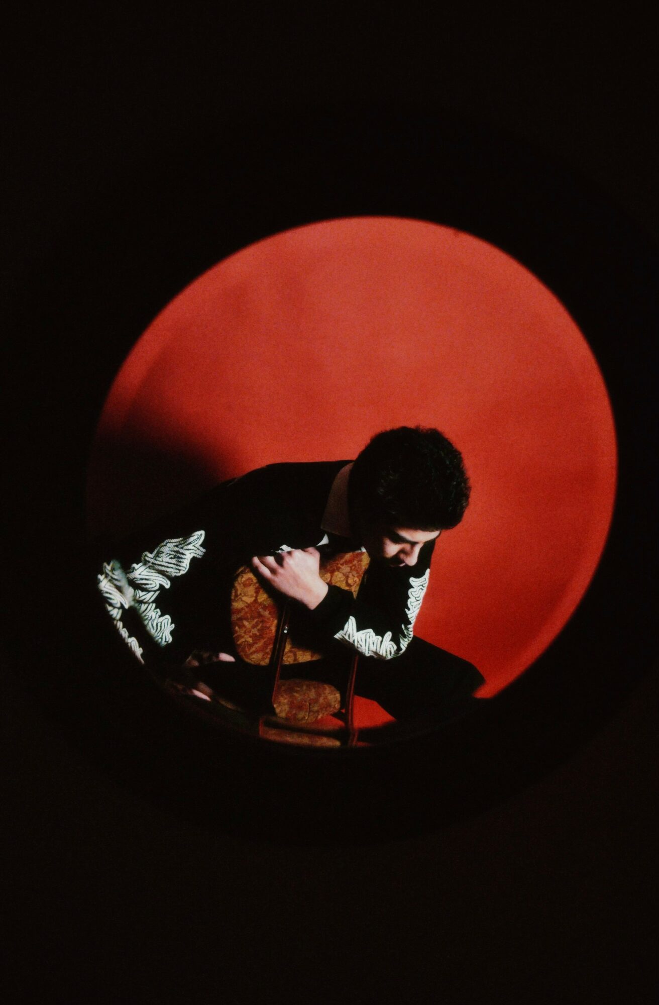 Person with curly hair and patterned clothing sitting in front of a large red circle, holding a camera, dark background