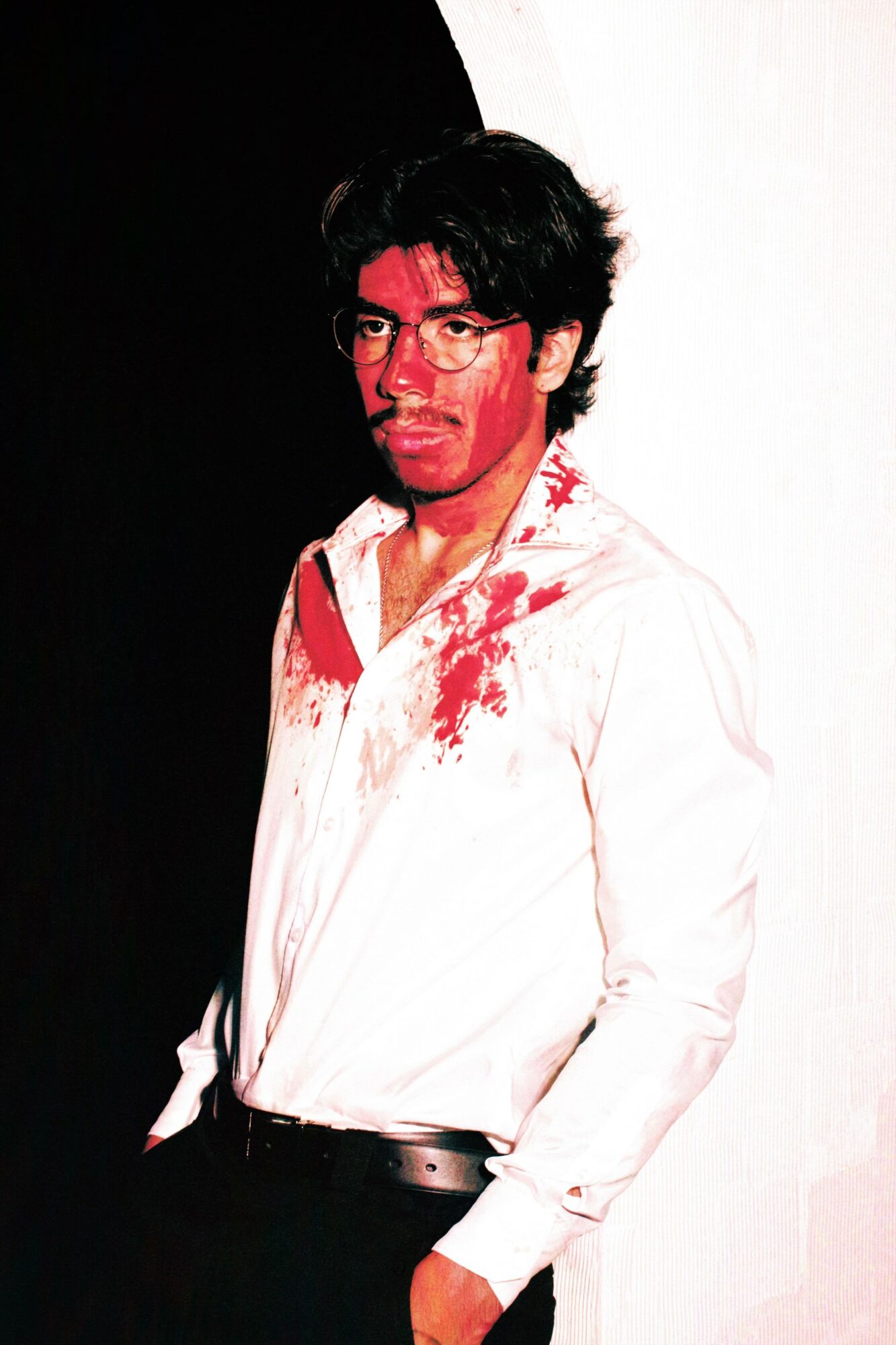 Person with glasses and dark hair, wearing a white shirt with red stains, standing against a black and white background.