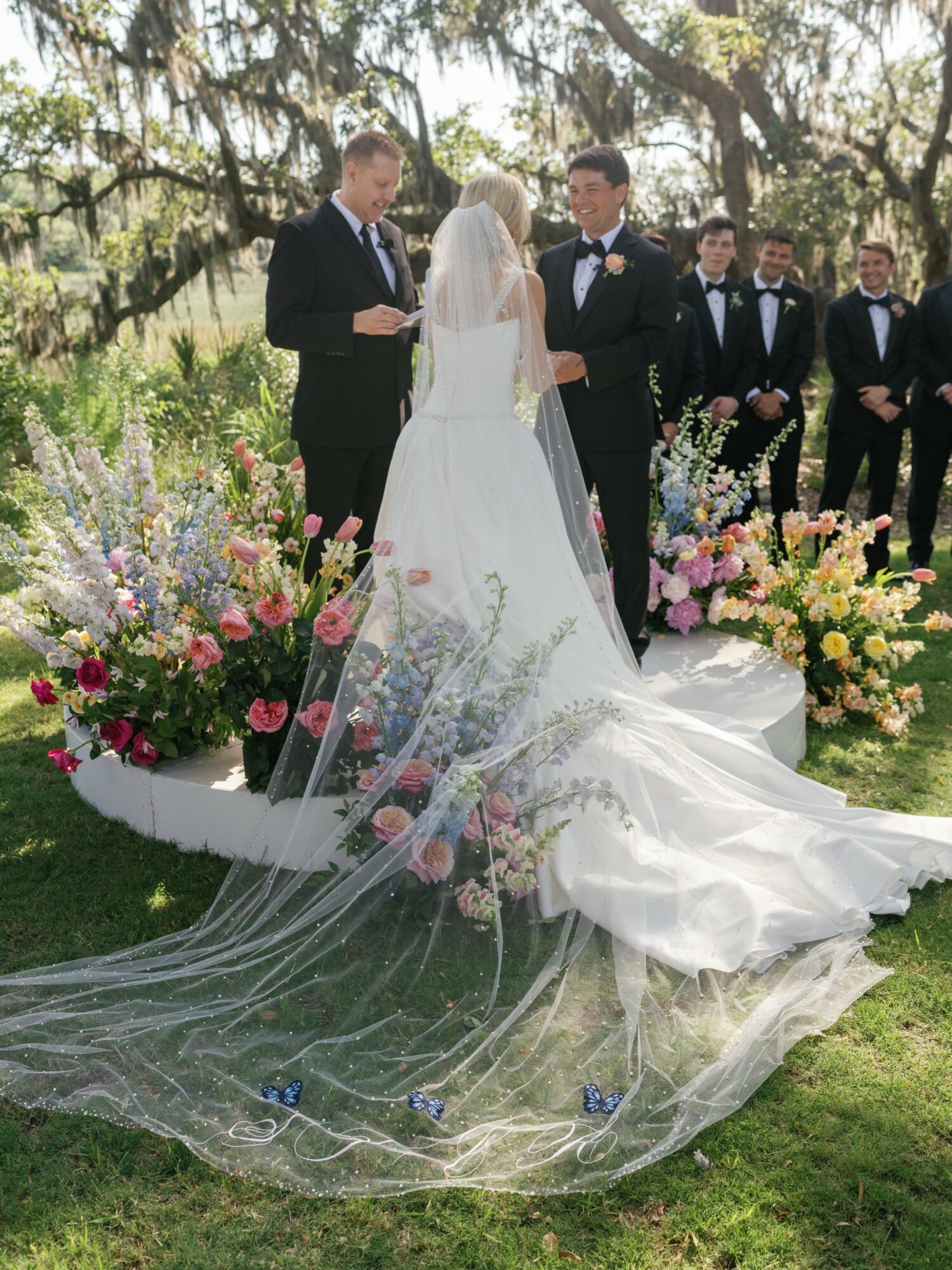 Bride and groom facing each other outdoors, surrounded by wedding party and floral arrangements, under trees.