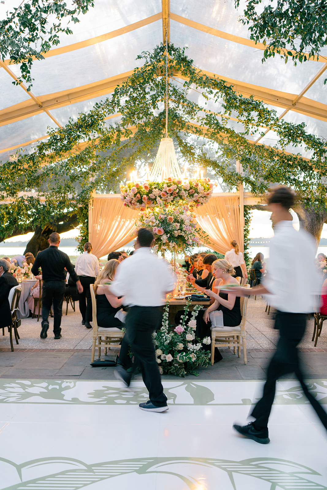 Indoor event with decorated ceiling, floral arrangements, and people seated and walking, sunlight streaming in from windows.