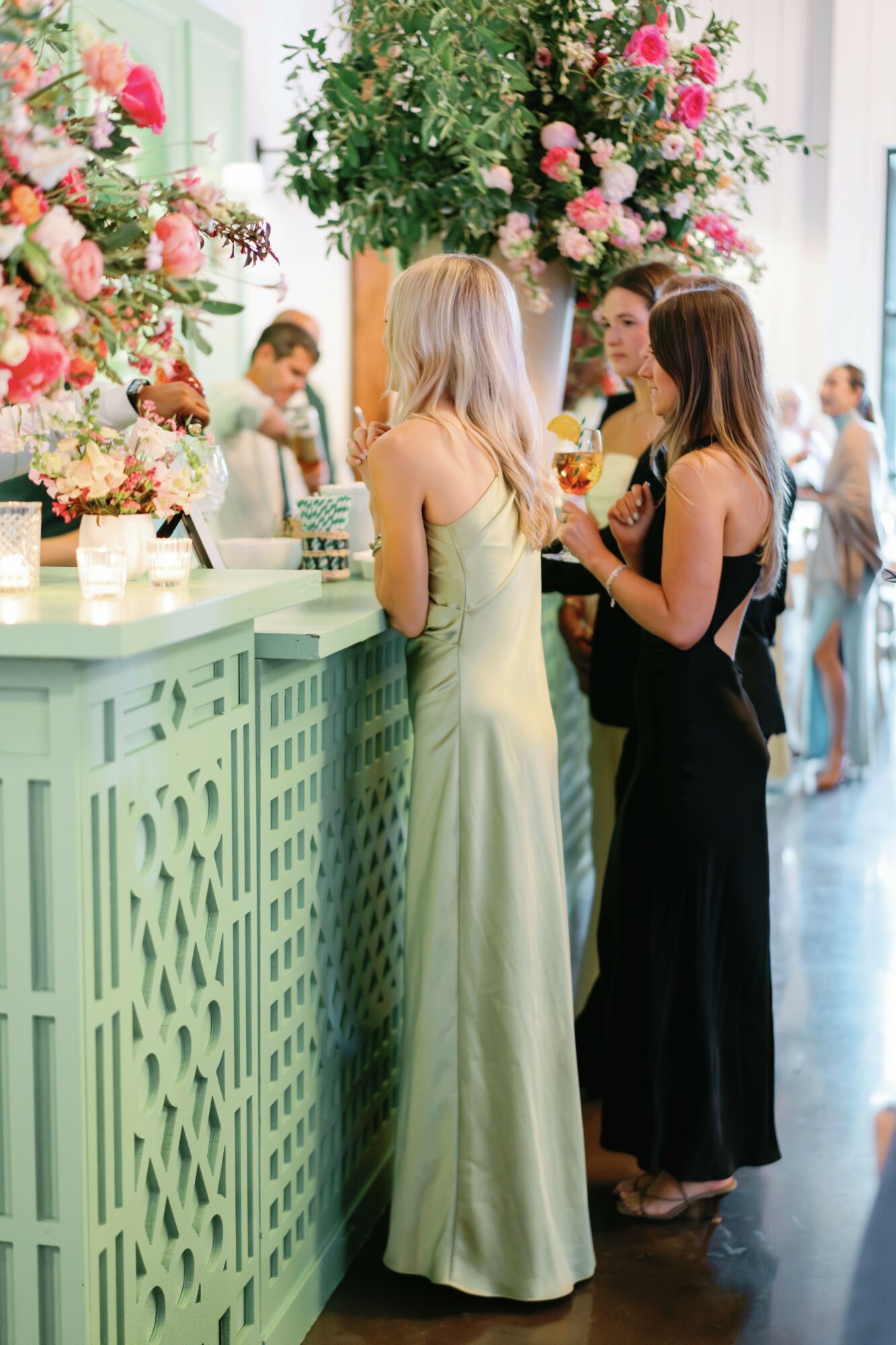 Two women in elegant dresses stand at a decorated reception desk with floral arrangements, engaging in conversation.
