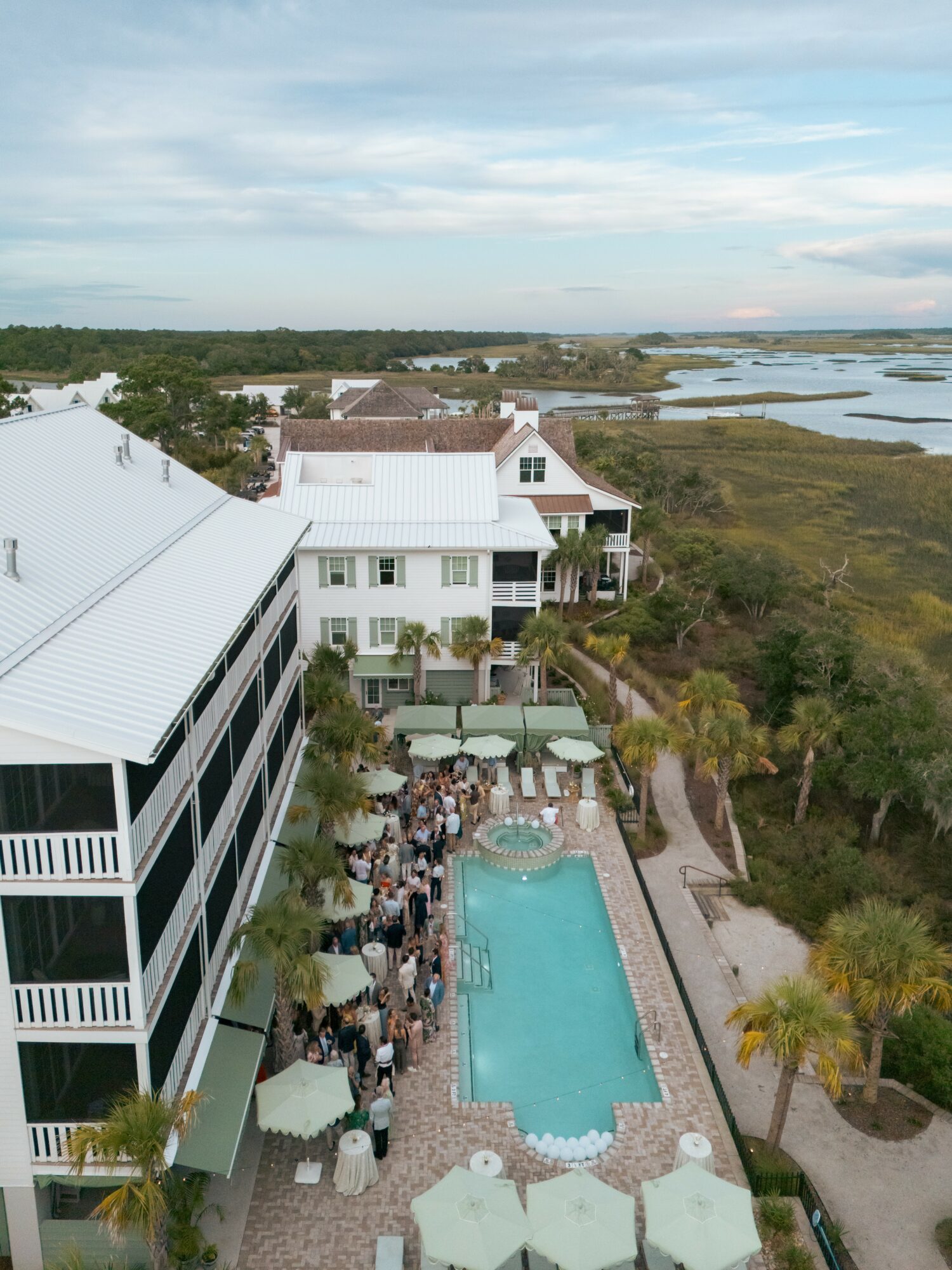 A swimming pool surrounded by people and umbrellas, with a building and marshland in the background.