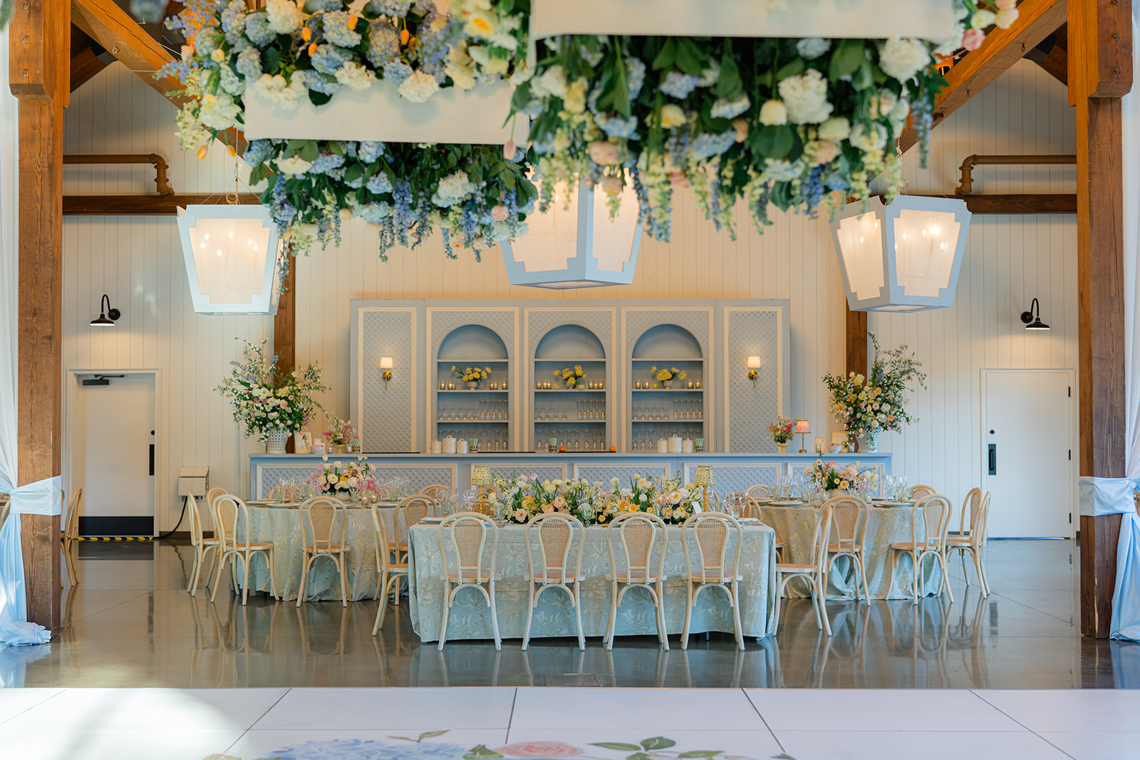 Elegant event space with a long table, floral arrangements, and a decorated ceiling, set in a bright room with wooden beams.