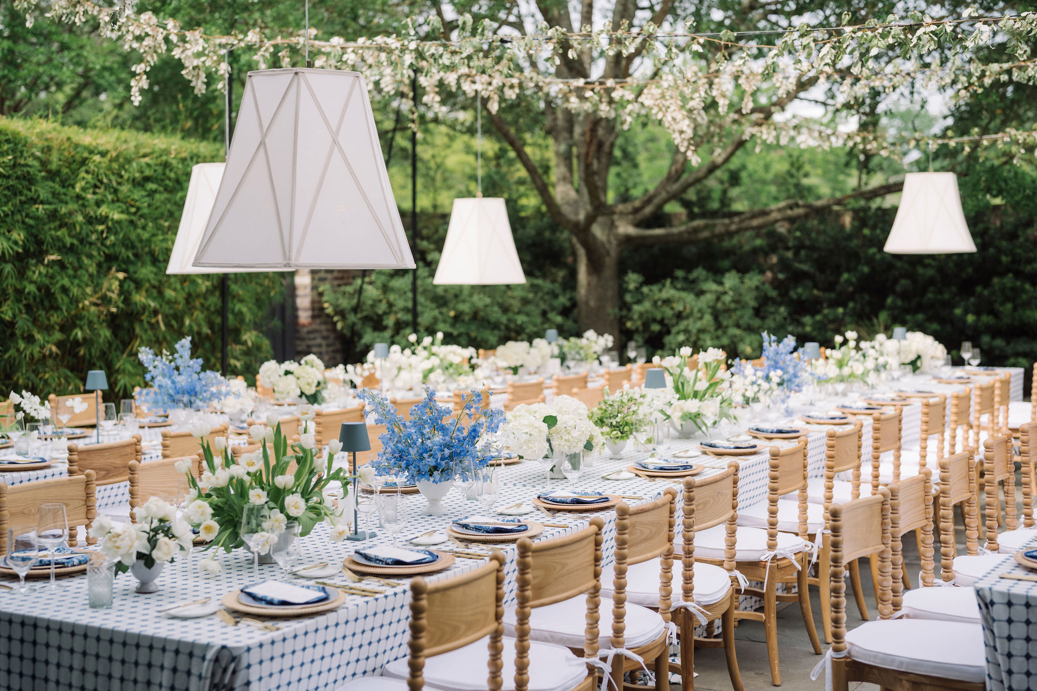 Long outdoor table set with white flowers, blue accents, and white lamps, surrounded by greenery and blooming trees.