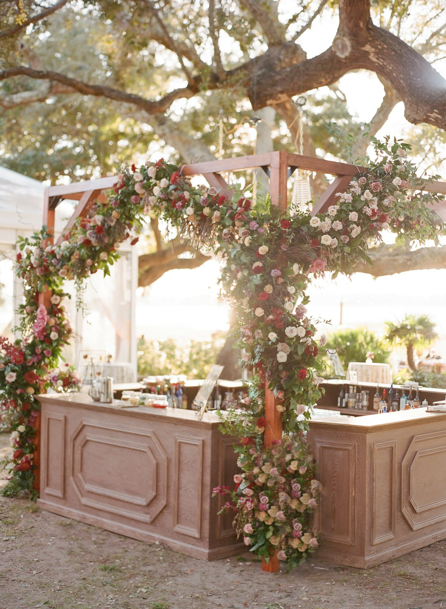 Wooden bar decorated with pink and white flowers, under a large tree with sunlight filtering through branches.