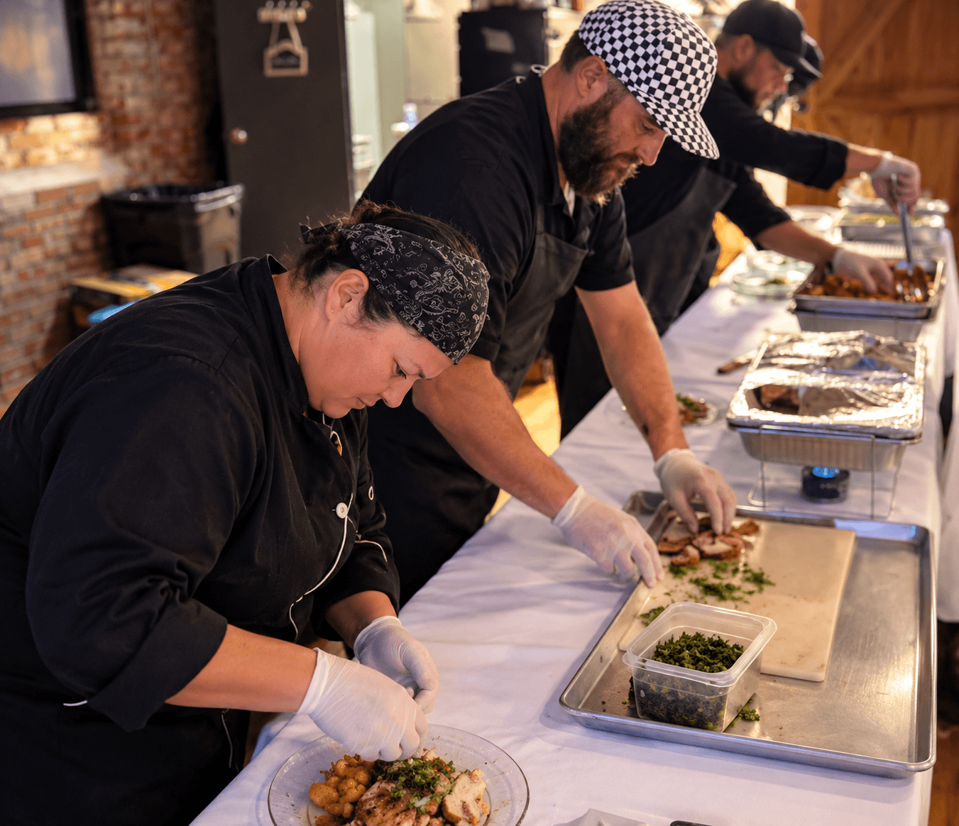 Three chefs preparing food at a long table with ingredients and trays, focused on their tasks in a kitchen setting.