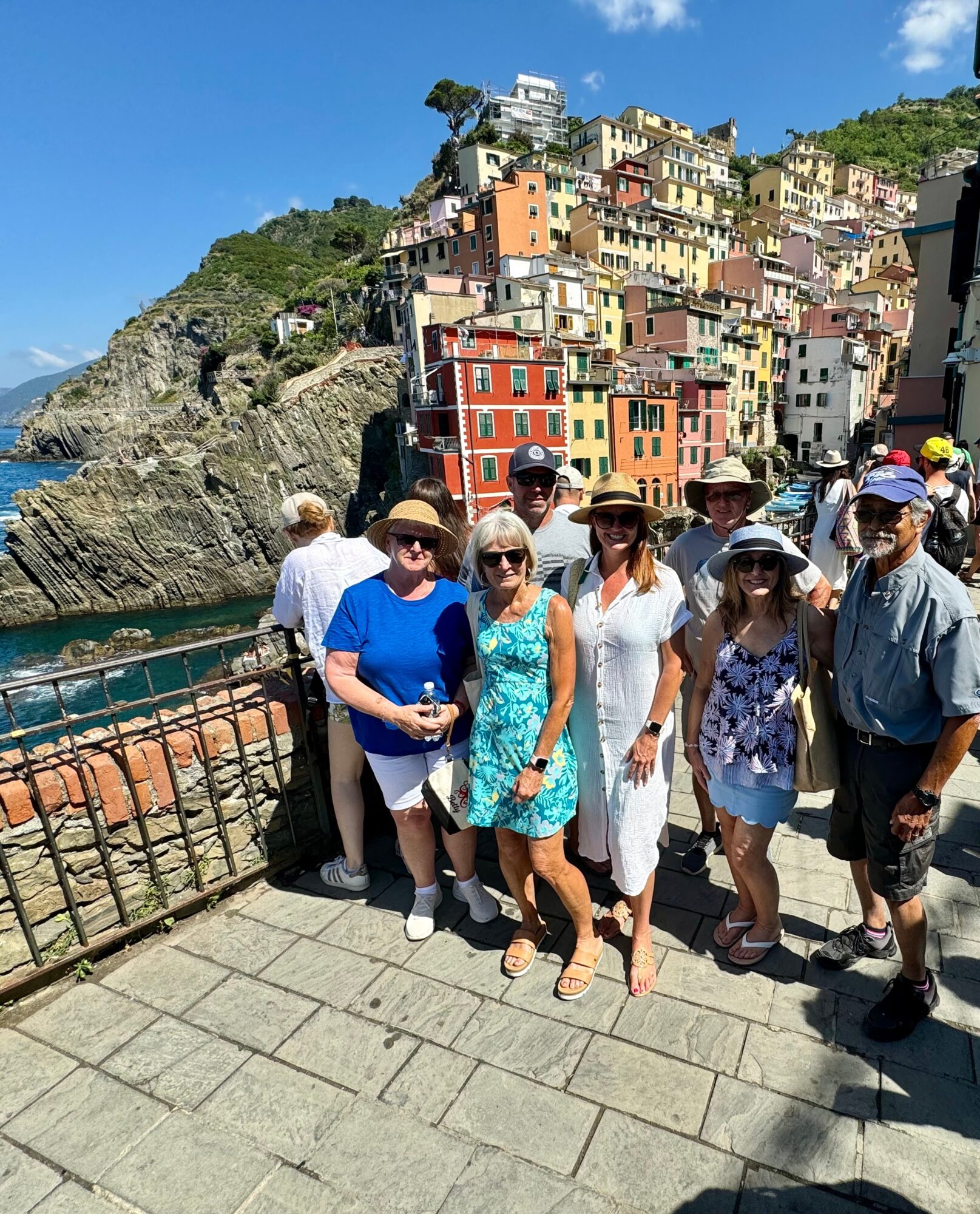 Five people pose on a sunny waterfront promenade below colorful buildings on a rocky hillside.