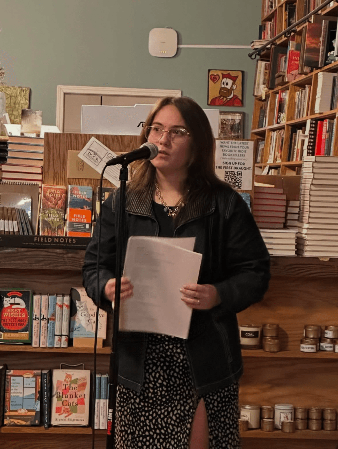 Young woman with glasses holding papers, speaking into a microphone in a bookstore or library setting.