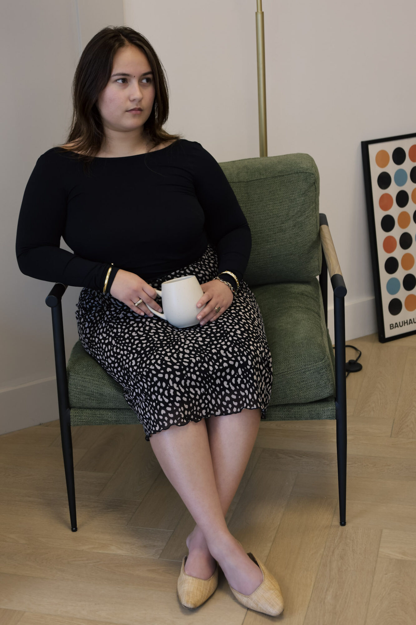 Young woman sitting on a green armchair holding a white mug, wearing a black top and patterned skirt, with a framed artwork nearby.