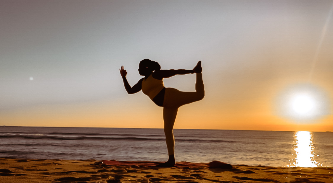 Person practicing yoga on beach at sunset, balancing on one leg with arm raised, ocean and sunset in background.