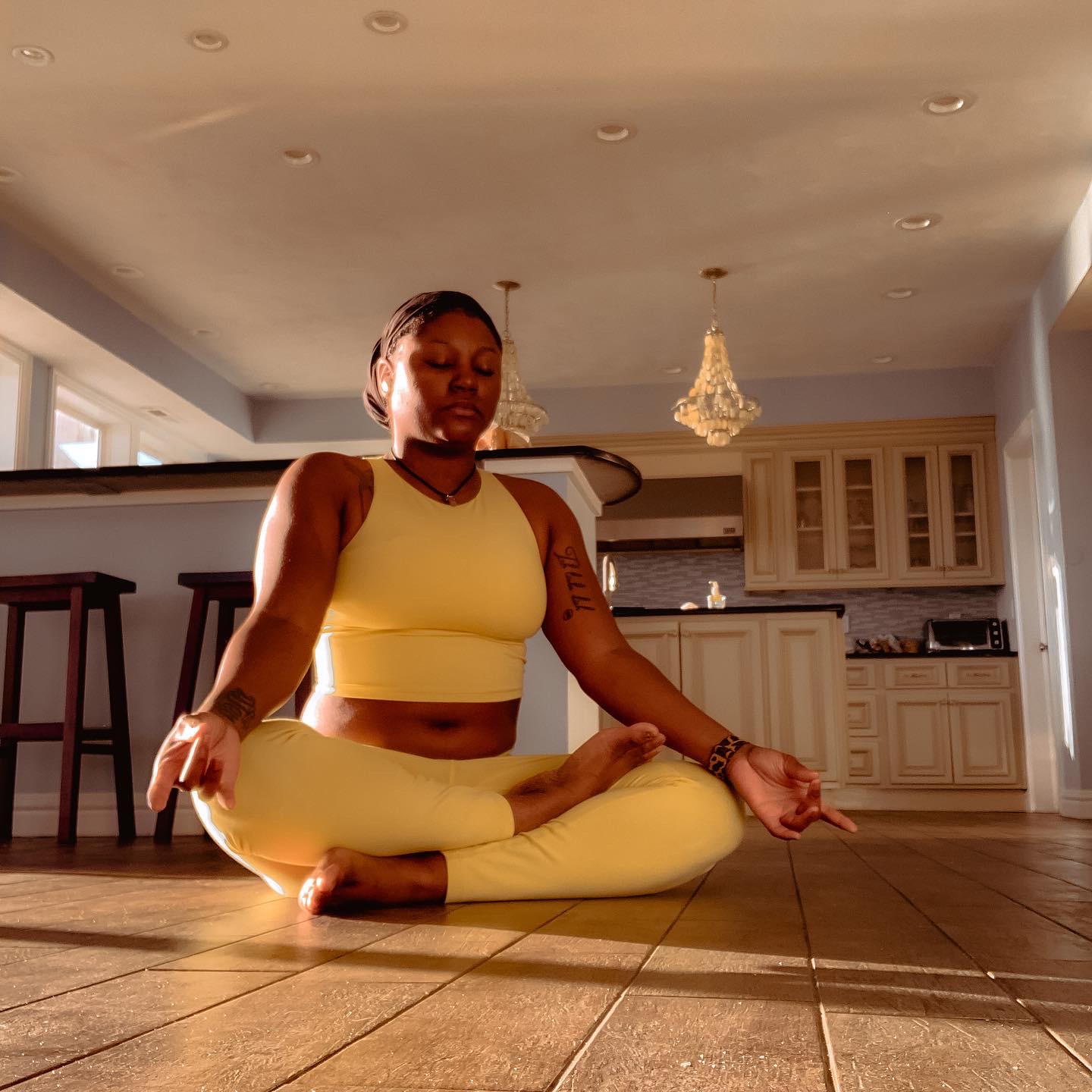 Person practicing yoga in a bright kitchen, sitting cross-legged with hands on knees, eyes closed, calm expression.