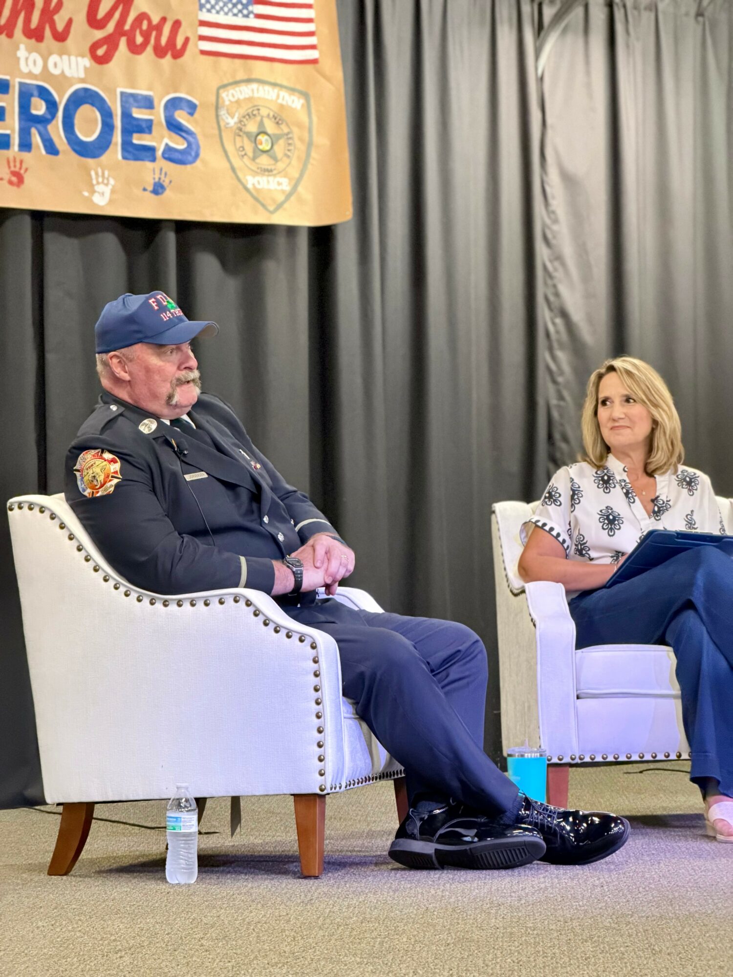 A man in a police uniform and a woman in a white blouse sit on white chairs on stage, with a patriotic banner behind them.