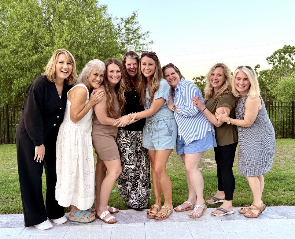 Group of eight women standing outdoors on grass, smiling, holding hands, with trees and a fence in background.