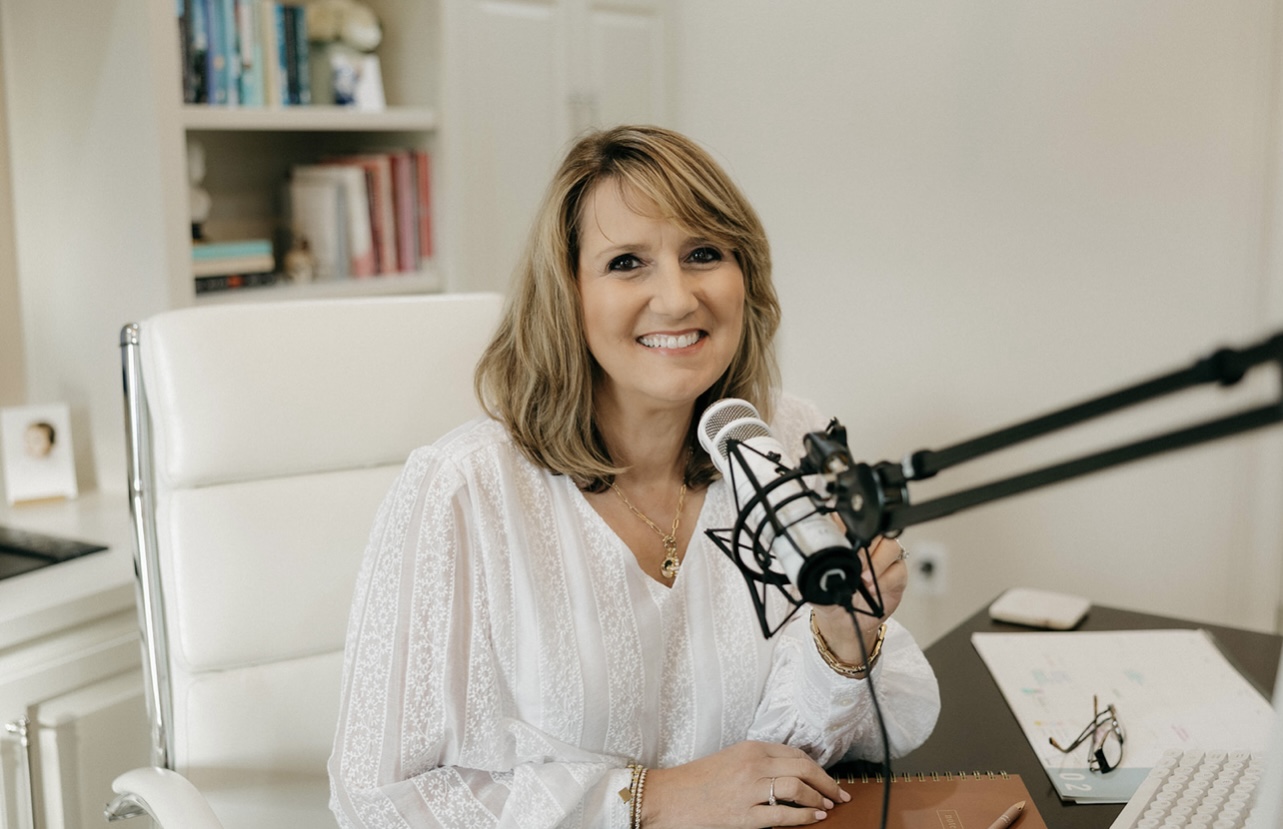 Woman smiling at microphone in a bright room with bookshelves, sitting at a desk with papers and a pen.