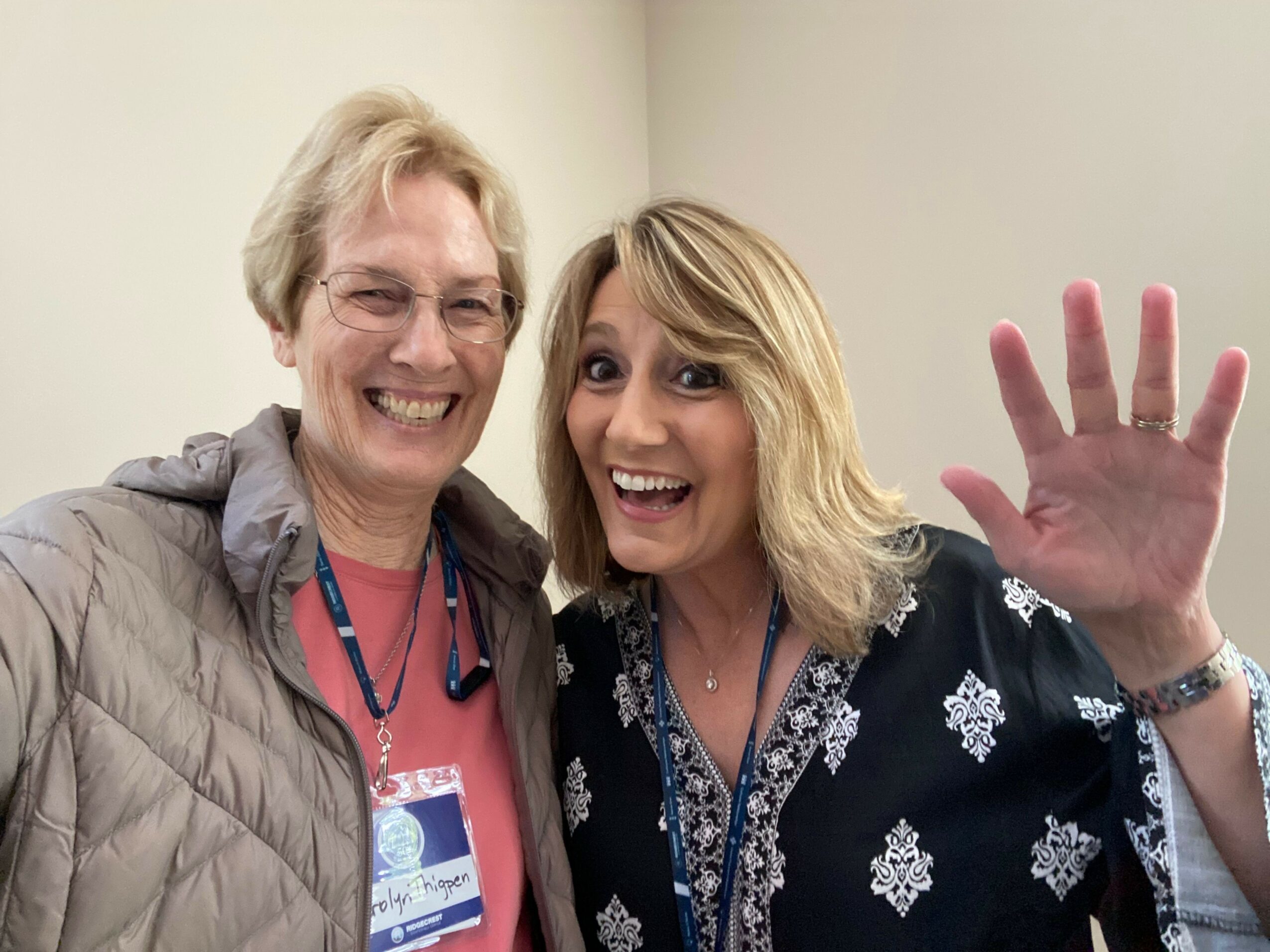 Two people smiling, one with blonde hair and the other with glasses, standing close together indoors.
