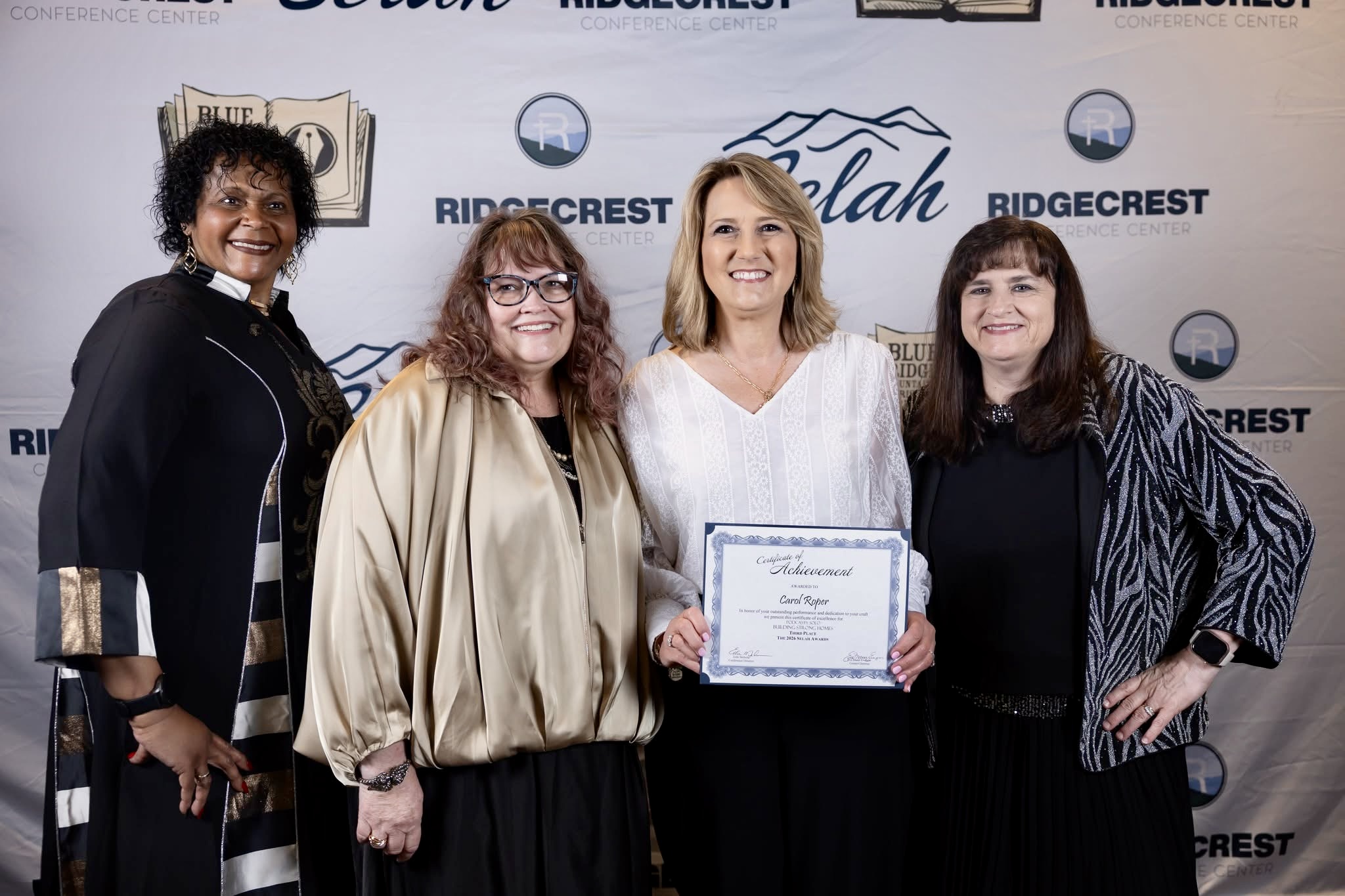 Four women standing together, one holding a certificate, smiling in front of a backdrop with logos.