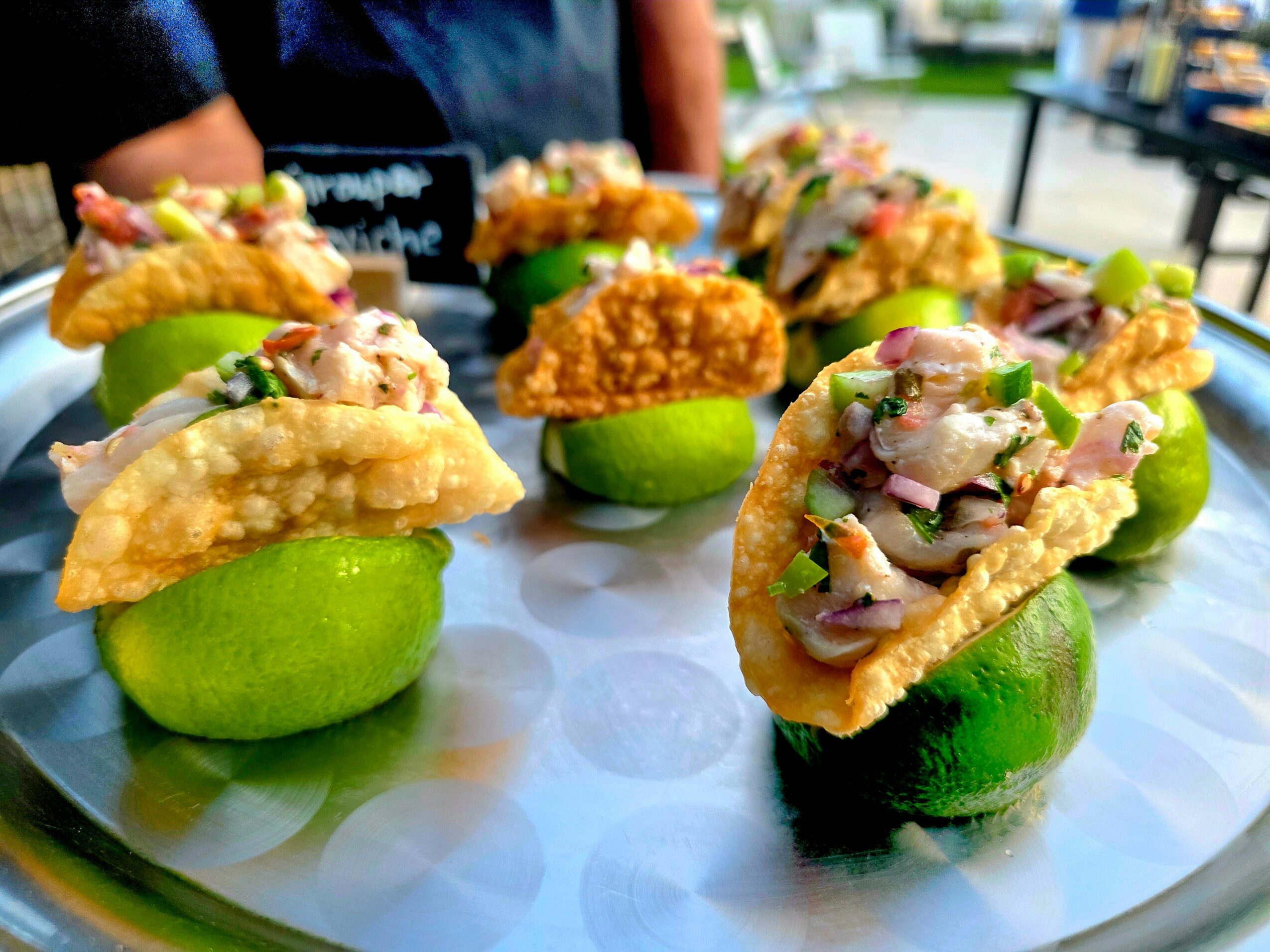 Several tacos with seafood and vegetables on lime wedges, arranged on a round tray outdoors.
