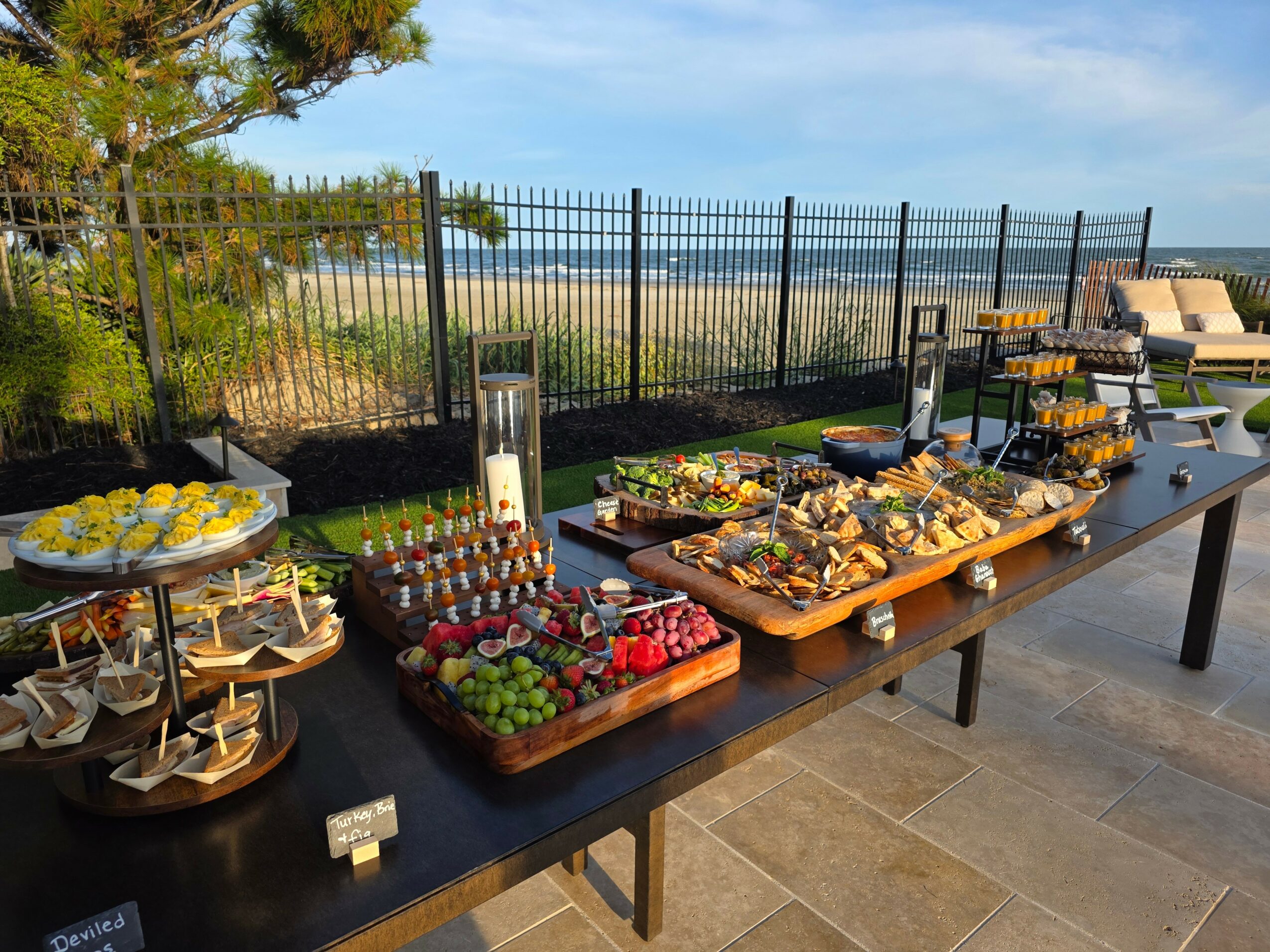 Outdoor buffet table with various food items, set against a fence and scenic landscape, under a blue sky.