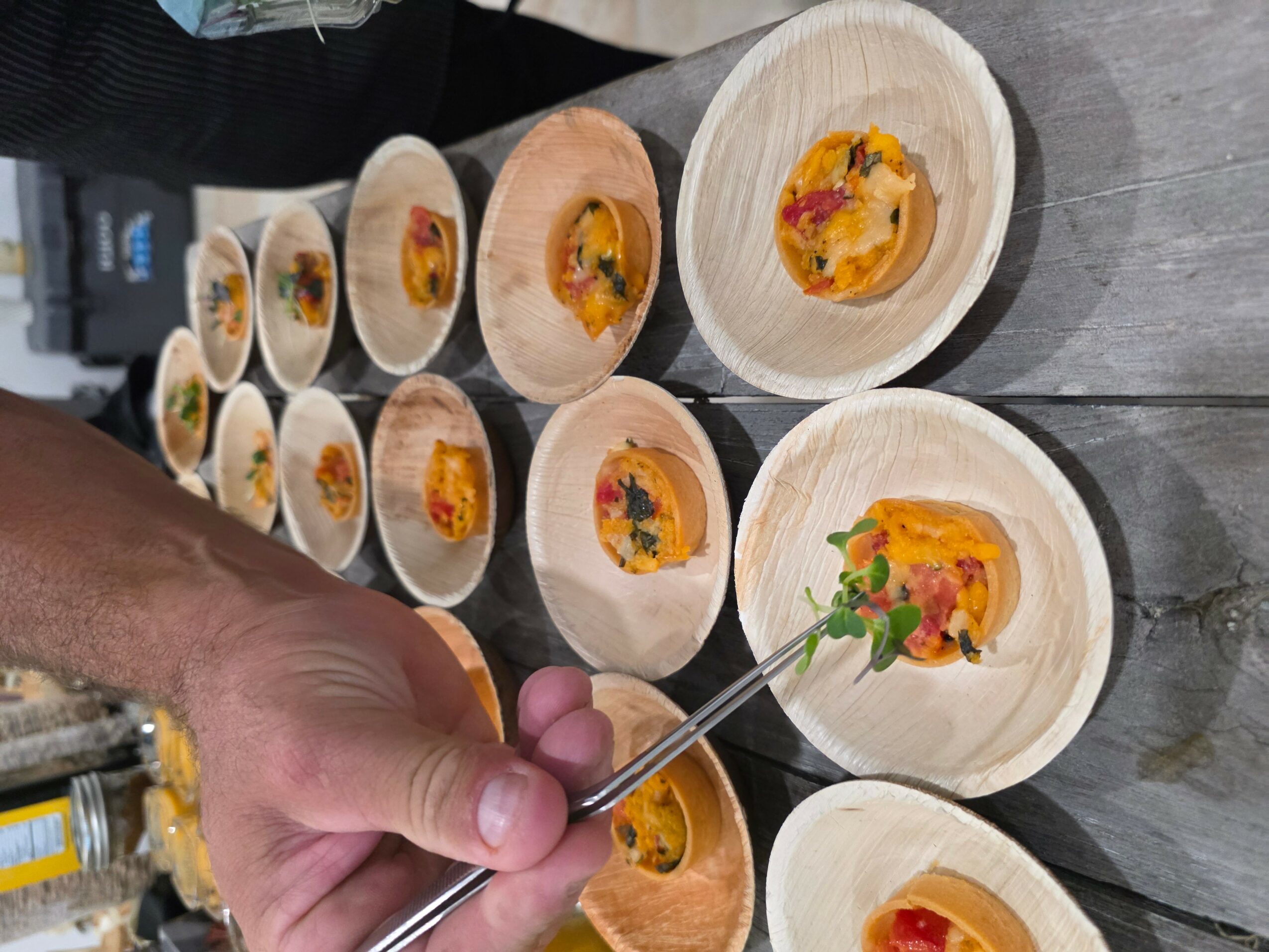 Person's hand holding a fork over small wooden bowls with colorful food, arranged on a wooden surface.