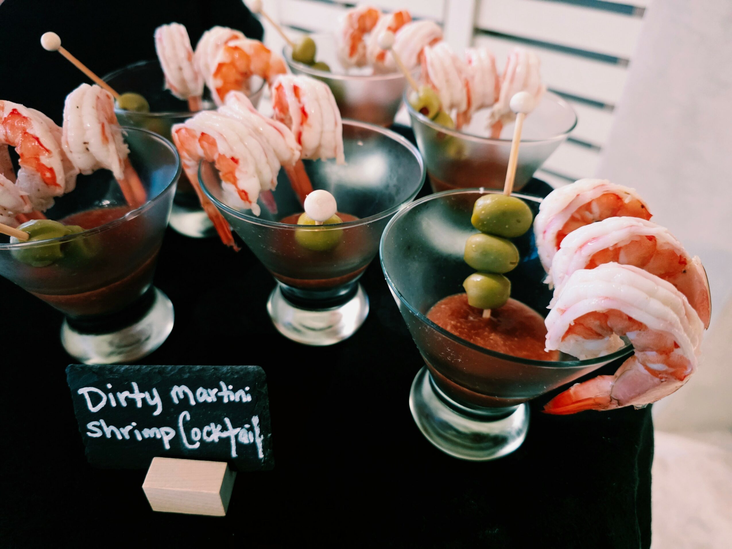 Four cocktail glasses with shrimp skewers and green olives on a black table, with a small chalkboard sign reading 'Dirty Martini Shrimp Cocktail'.