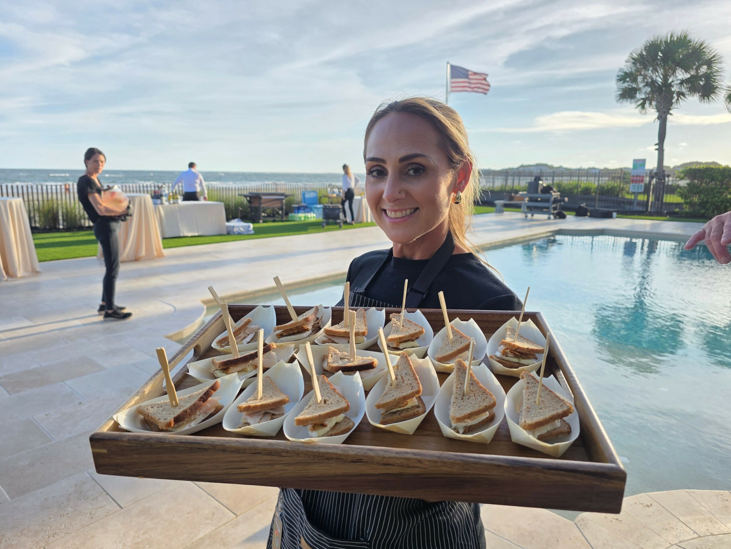 Woman holding a tray of appetizers near a pool with an American flag and palm trees in the background.