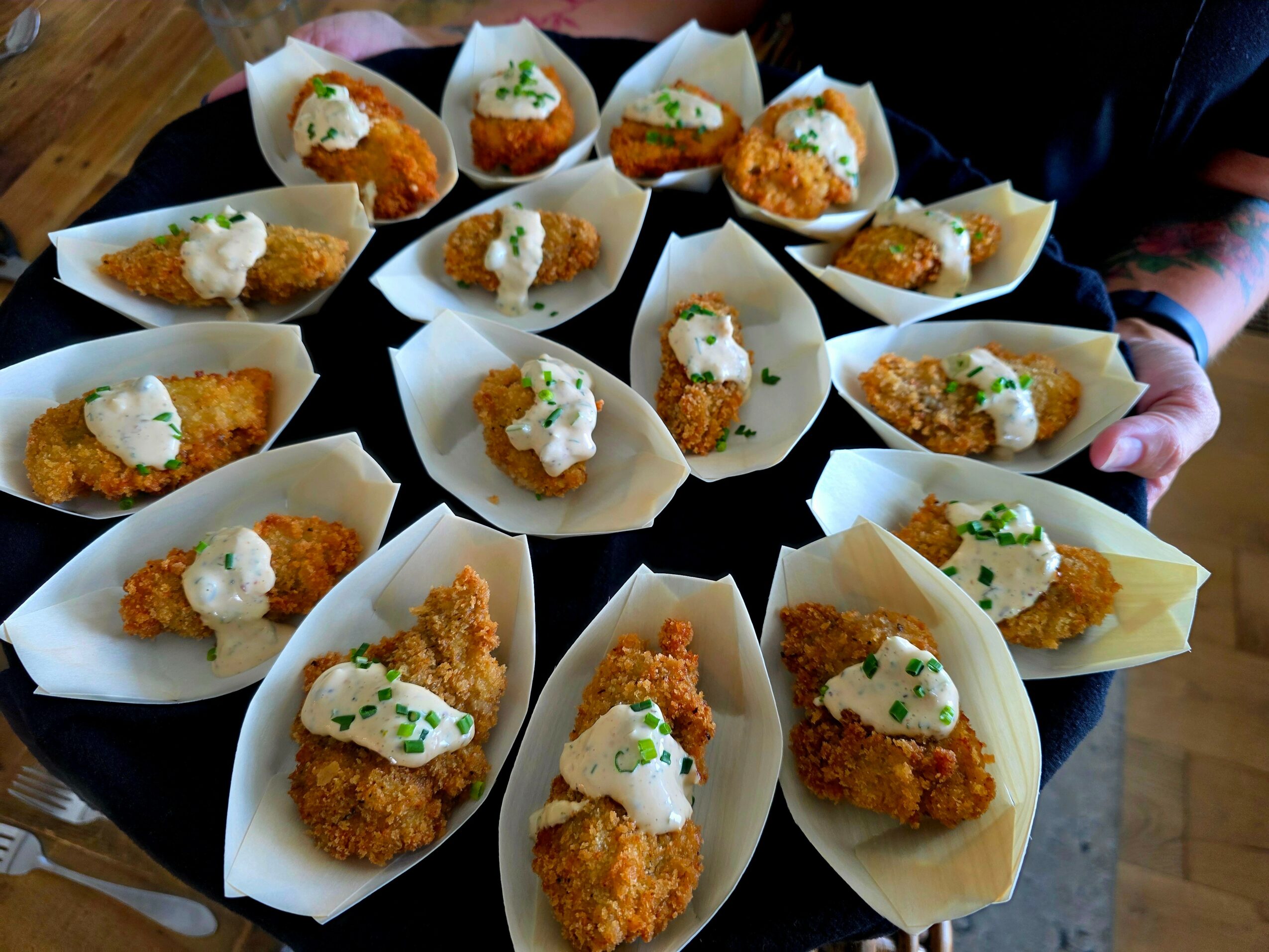 Multiple fried food pieces topped with sauce and green herbs, served in paper boats on a black tray.