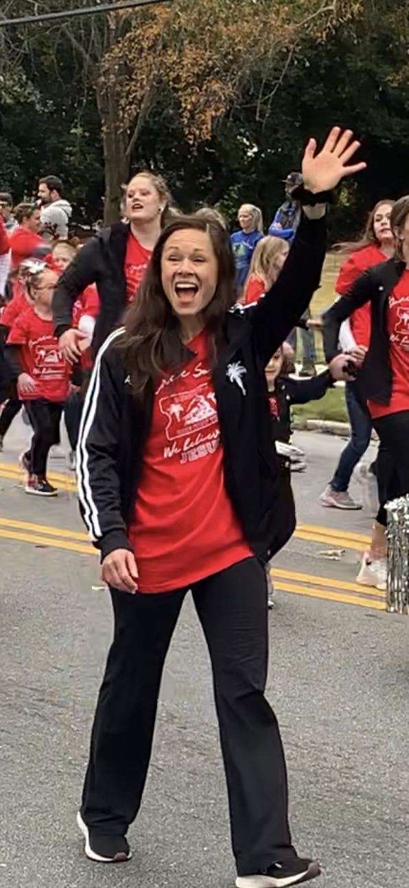 A person in a red shirt waves beside a parade route as a crowd walks behind them.