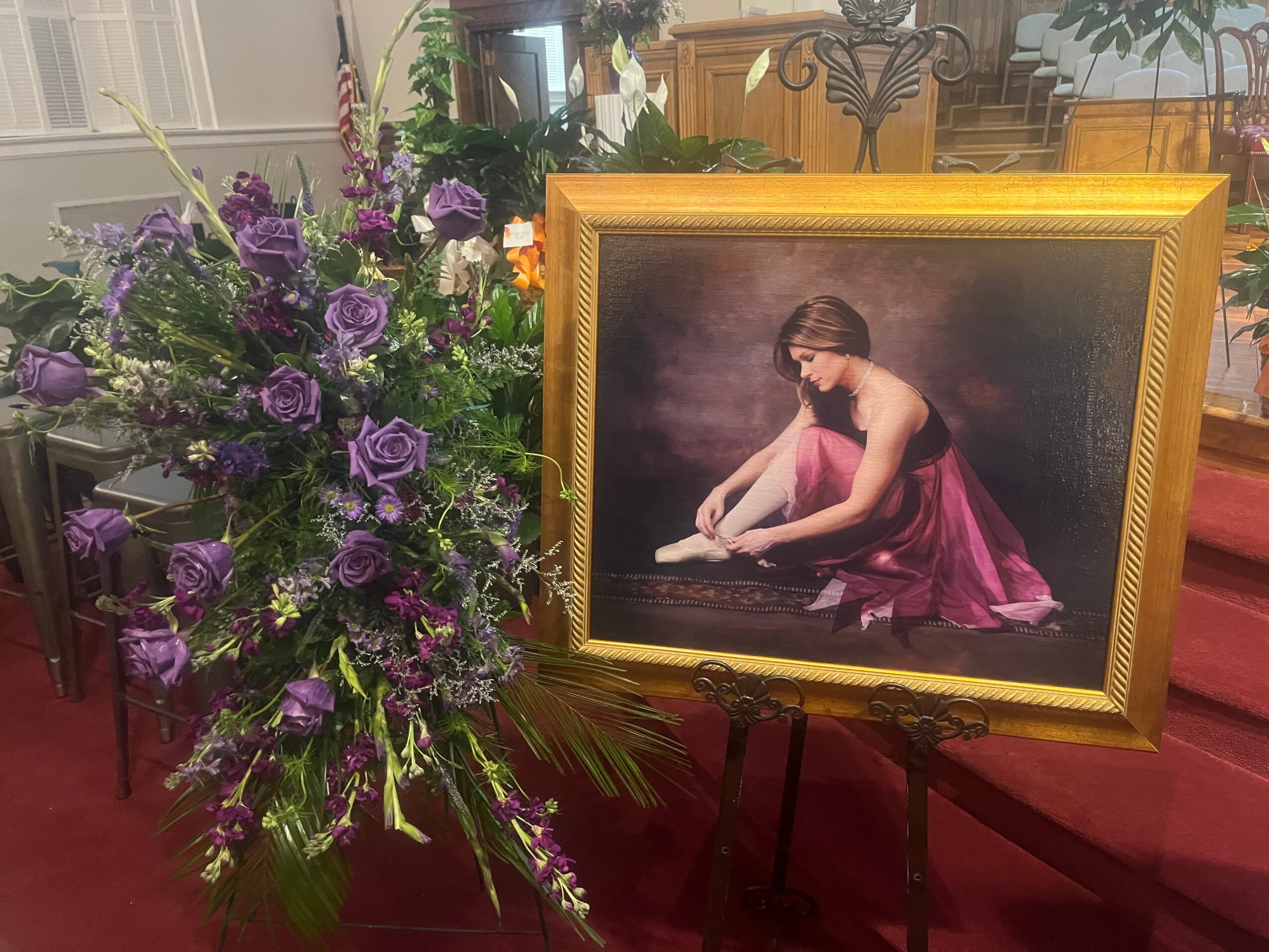 A framed painting of a seated woman beside a large arrangement of purple flowers in a room.