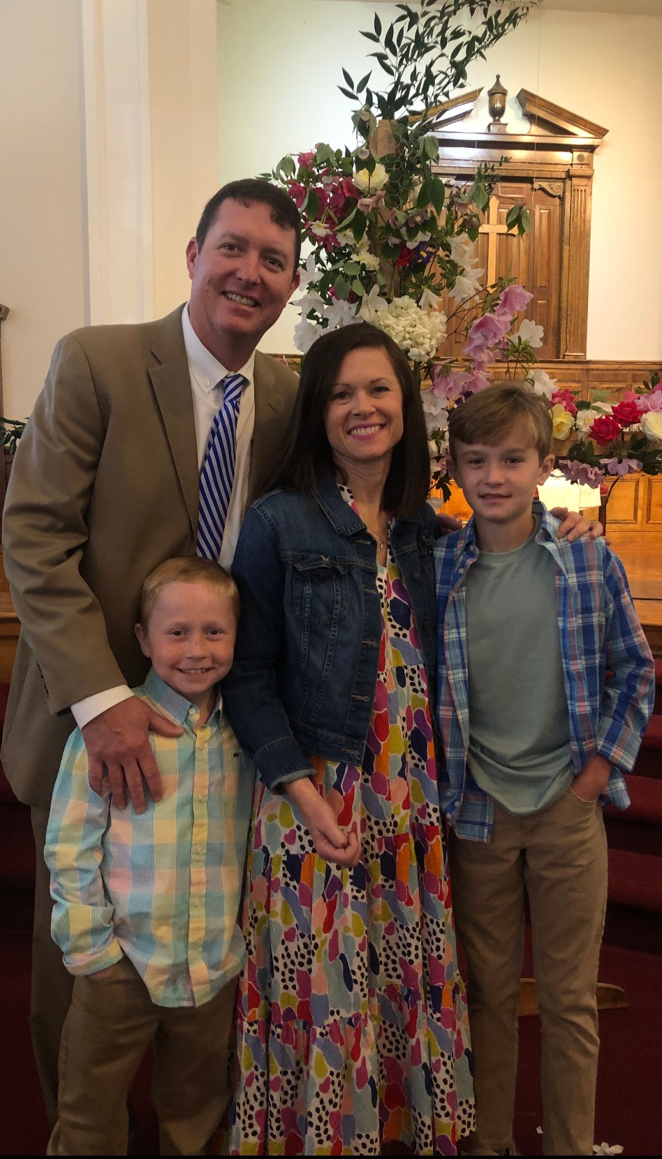 Four people stand together in a decorated room with flowers and a wooden cabinet.