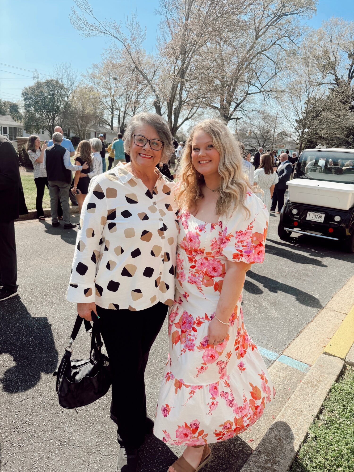 Two people stand together outdoors near parked vehicles and leafless trees.