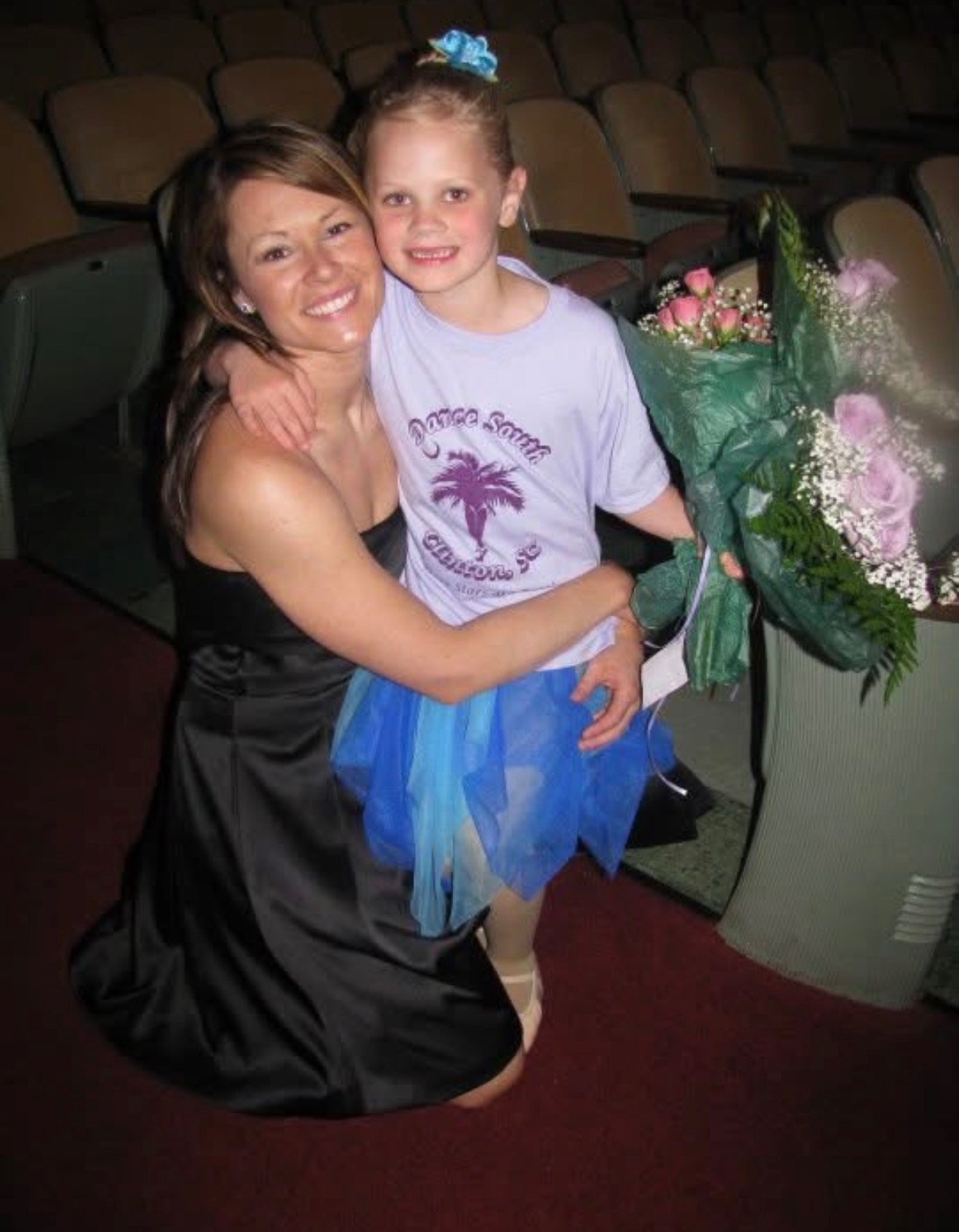 A woman holds a child in a blue skirt beside a large bouquet in an auditorium.