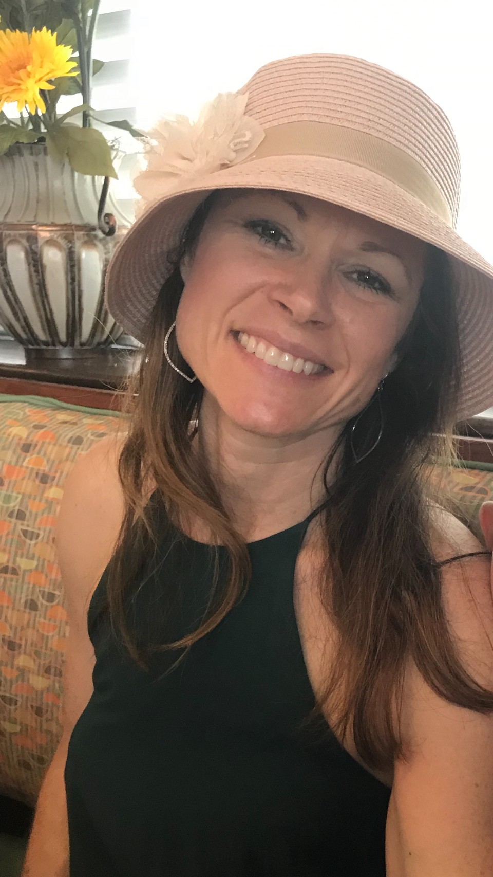 A smiling person wearing a wide-brimmed straw hat and dark sleeveless top, seated beside a vase of flowers.