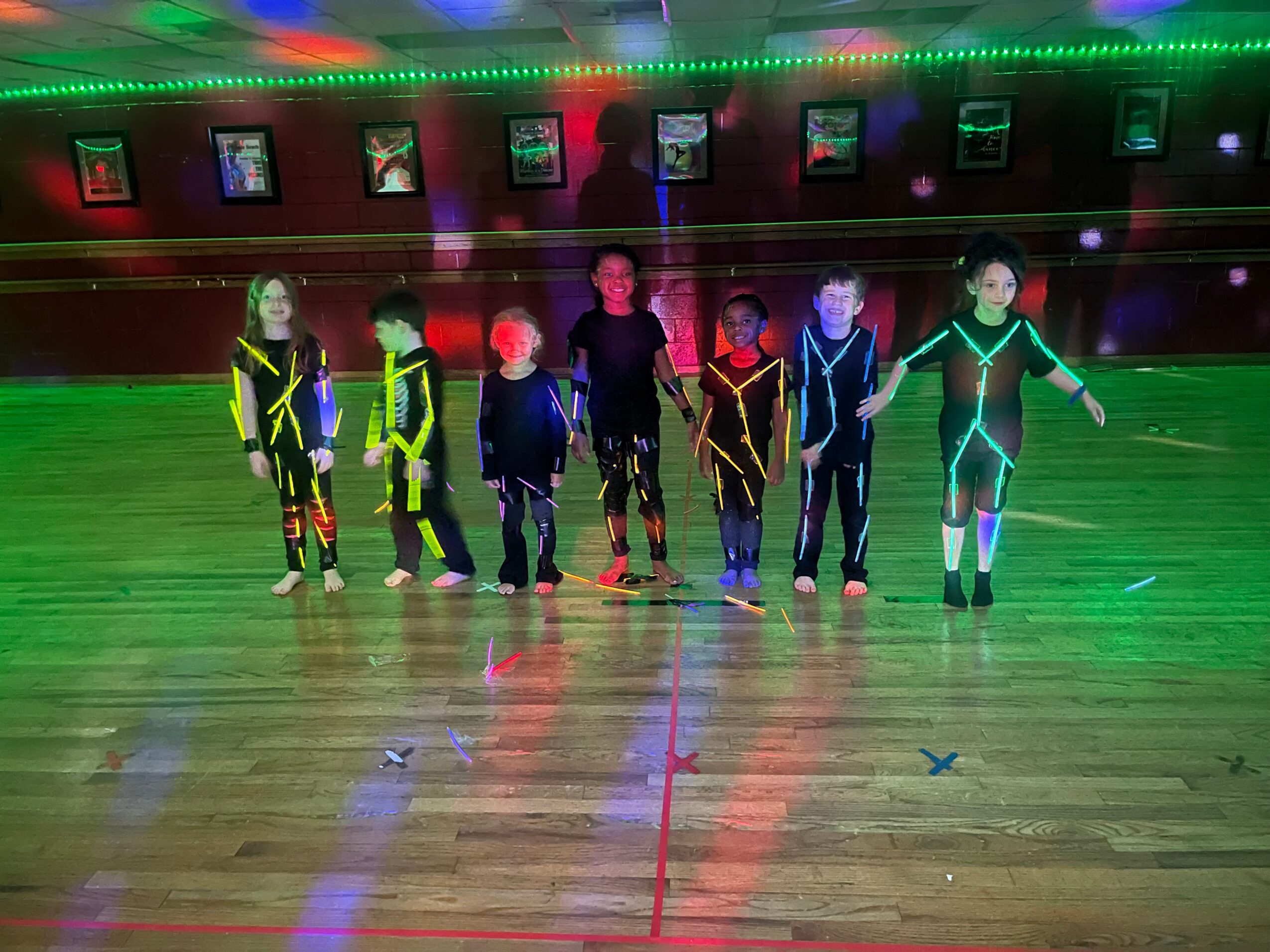 A group of children in colorful clothing stands on a polished floor beneath green and red lights.
