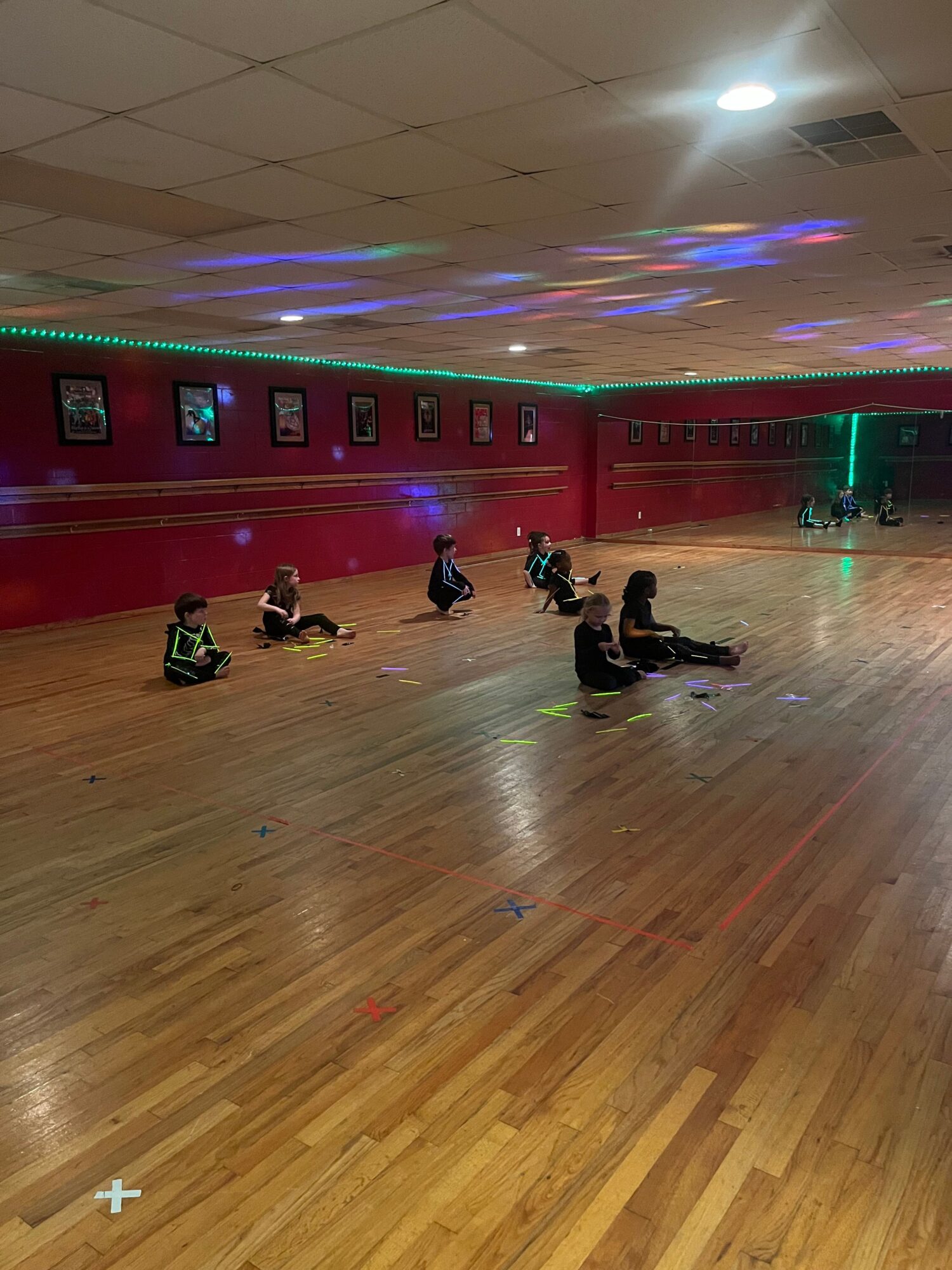 People sit along the edge of a wooden floor beneath colorful lights in a gymnasium.
