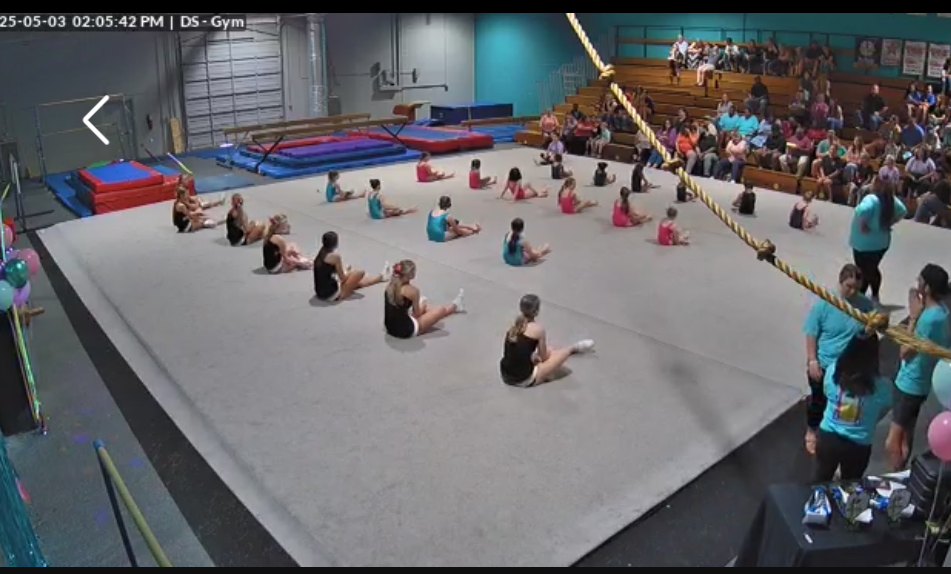 People sit on a large padded gym floor while others watch from the side.