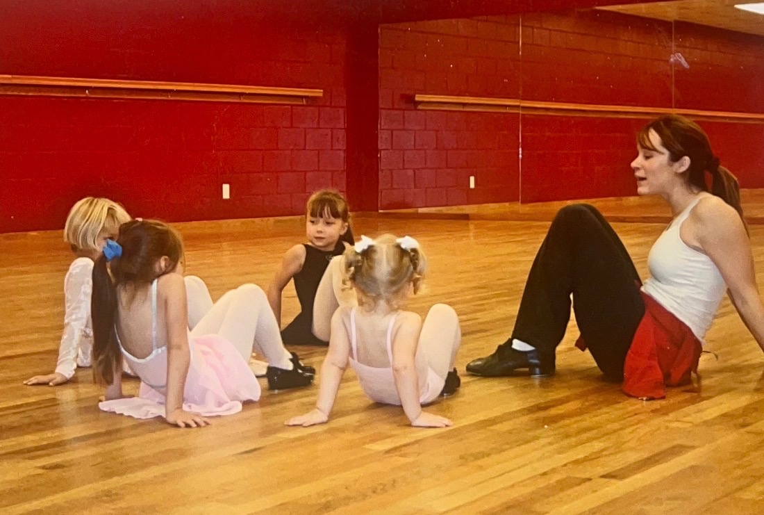 Four people sit on a wooden floor in a dance studio with red walls.