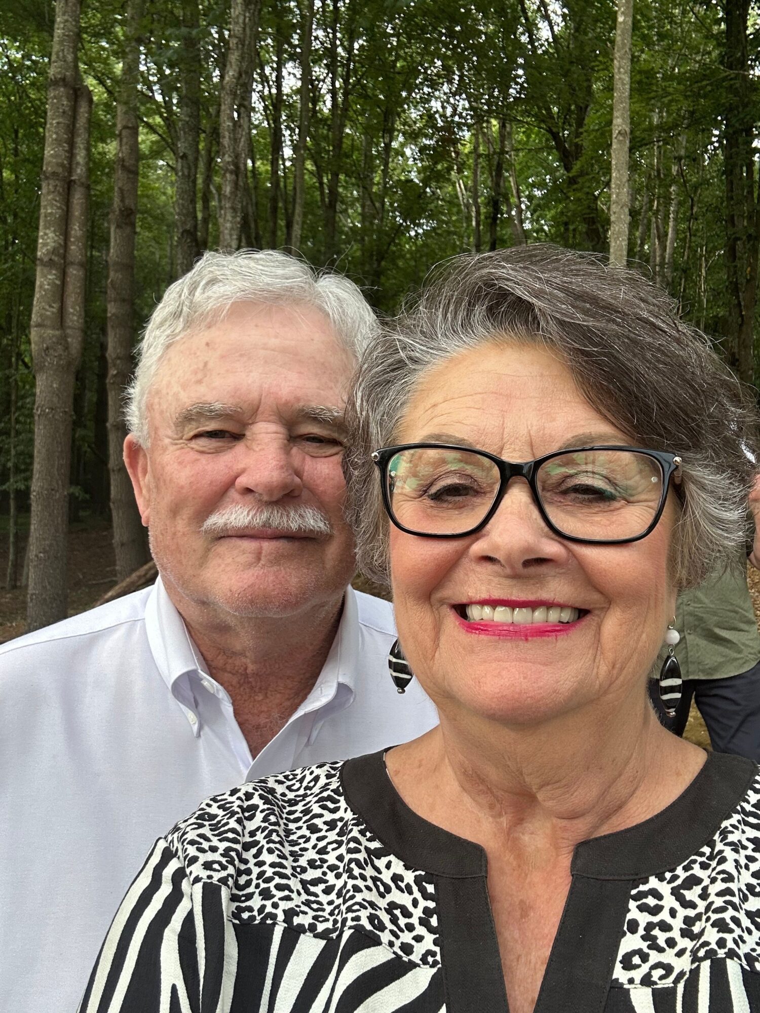 Two people pose outdoors in front of trees; one wears glasses and a patterned top, the other a white shirt.